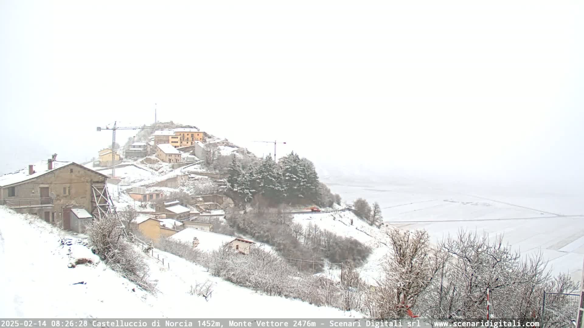 A snow-covered village nestled on a hillside, with construction cranes visible, overlooks a flat, snow-covered plain under a hazy, snowy sky.