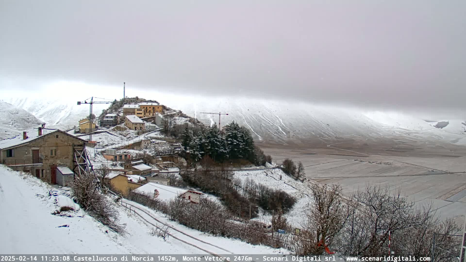 A snow-covered village nestled on a hillside, with a construction crane visible, overlooks a flat, snow-dusted plain under a cloudy, overcast sky.