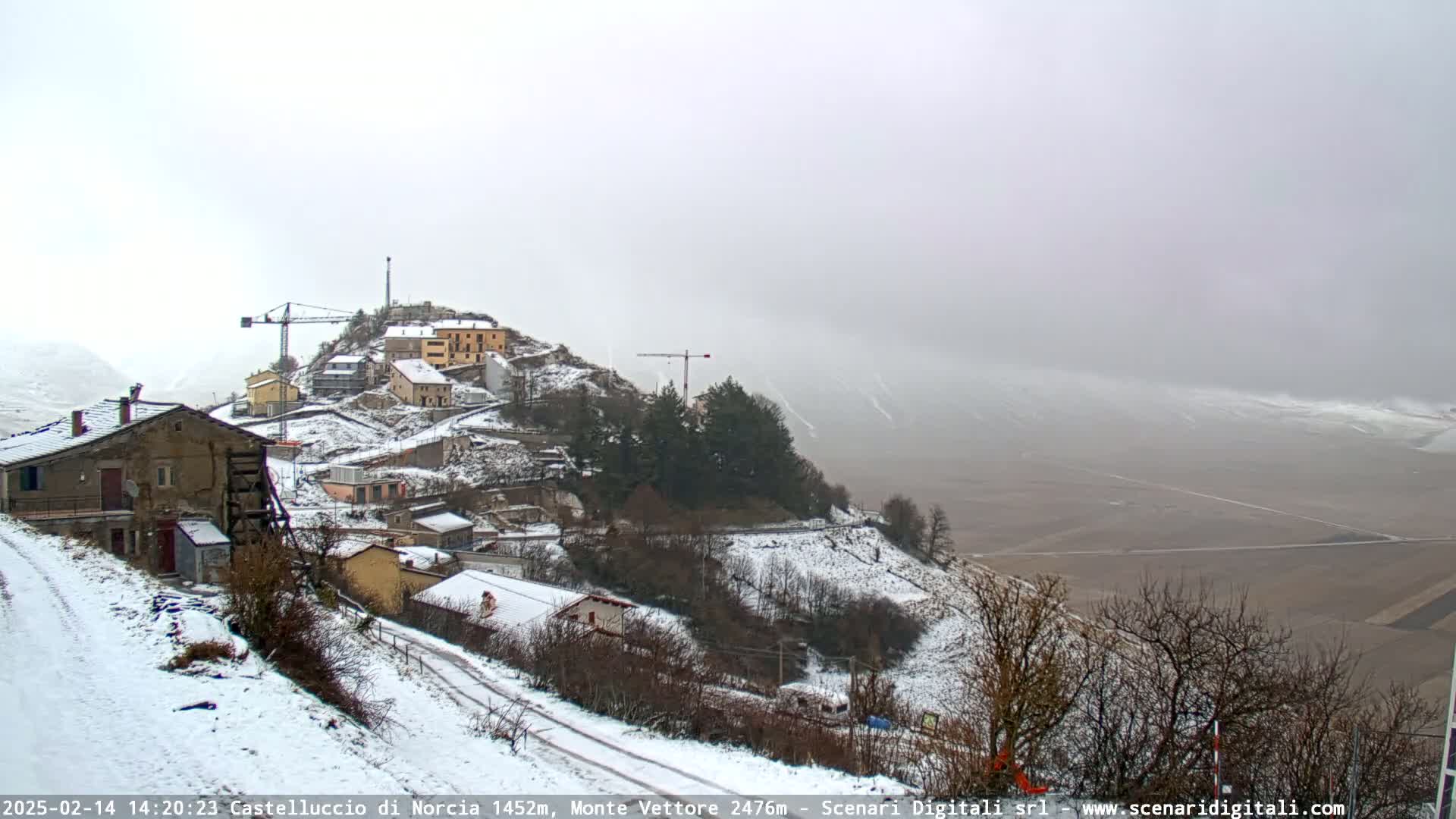 A snow-covered village nestled on a hillside, with construction cranes visible, under a hazy, overcast sky.