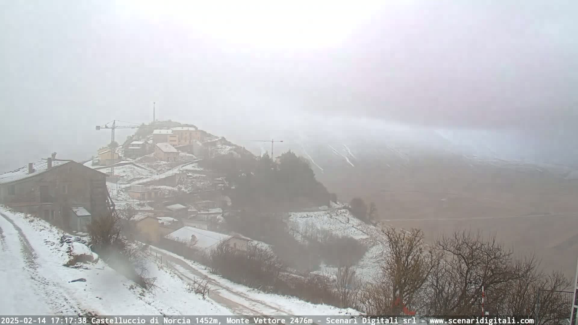 A snow-covered village nestled on a hillside, with a construction crane visible, is shrouded in a thick fog under snowy conditions.