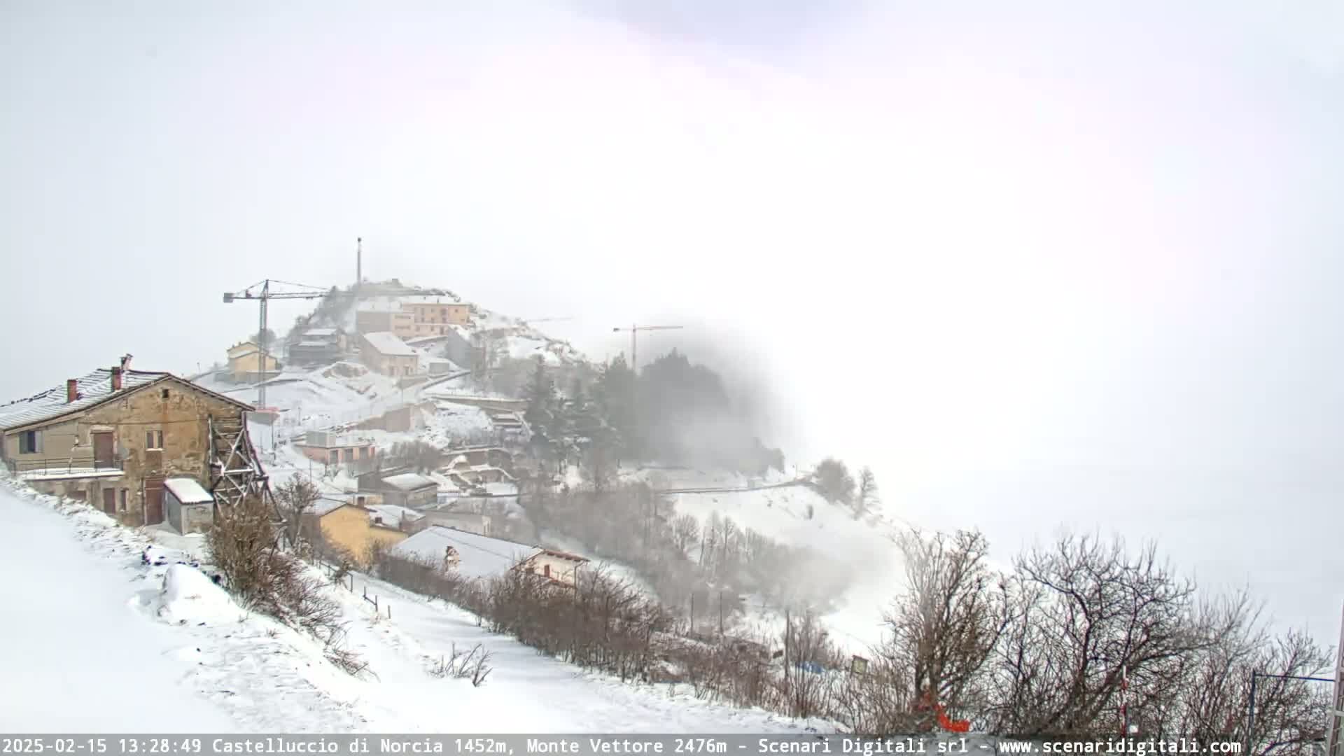 A snow-covered hillside village with construction cranes is shrouded in fog under an overcast sky.
