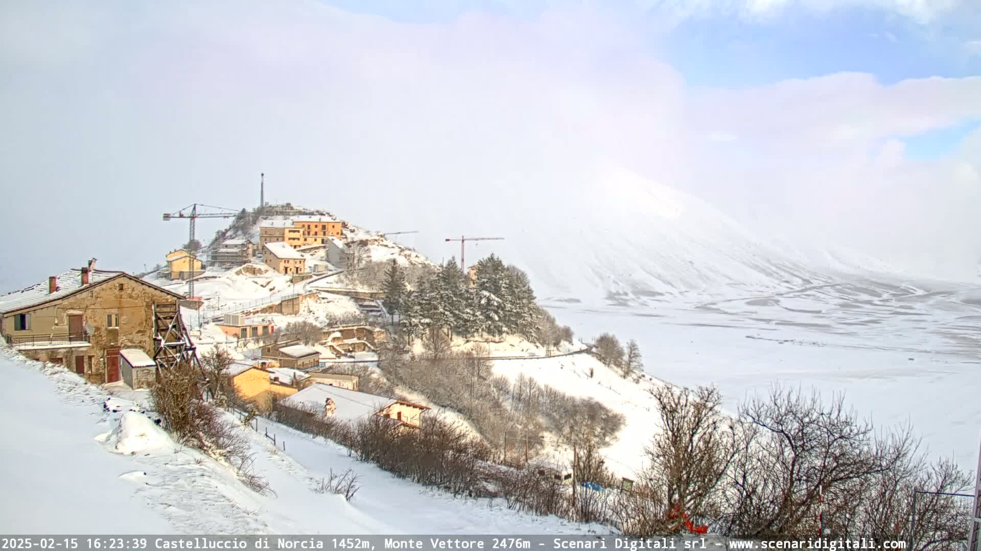 A snow-covered village nestled on a hillside, with construction cranes visible, overlooks a vast, snowy plain under a partly cloudy sky.