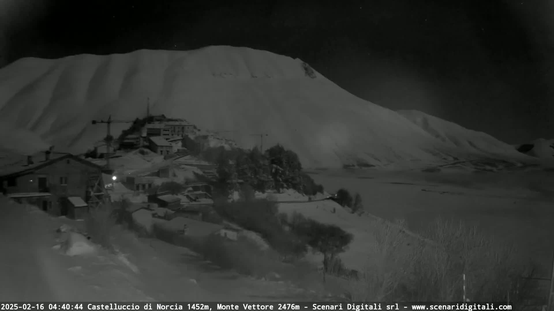 A snow-covered village is nestled at the foot of a large, snow-dusted mountain under a dark, nighttime sky.