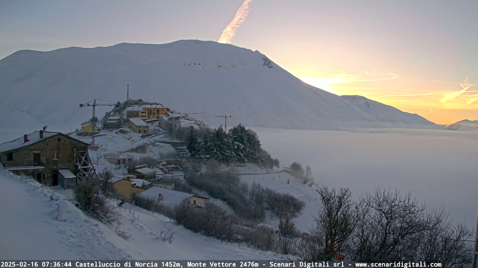 A snow-covered village nestled in a valley below a large, snow-capped mountain at sunrise, with a layer of fog or low clouds in the valley.