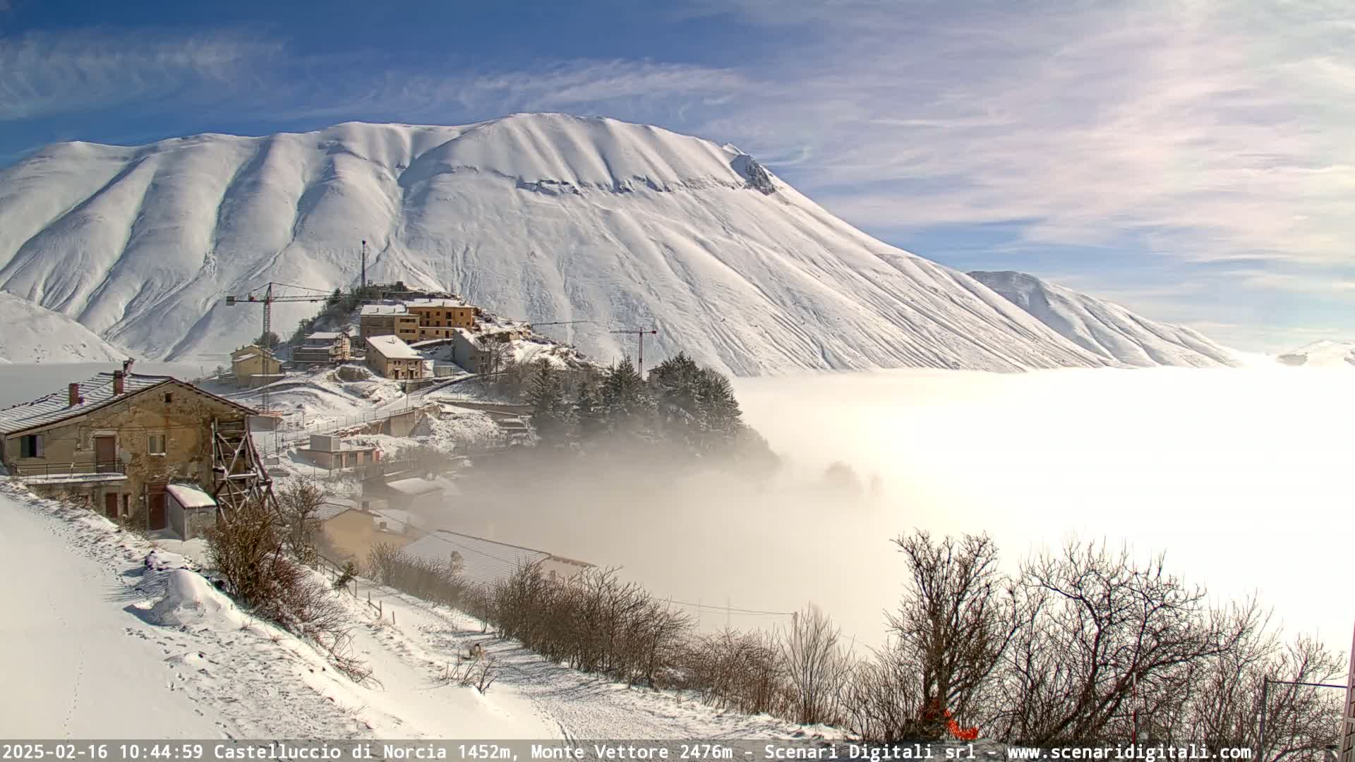 A snow-covered village nestled in a valley below a large, snow-capped mountain, with a layer of fog or low clouds partially obscuring the valley and partially covering the village, under a mostly sunny sky.