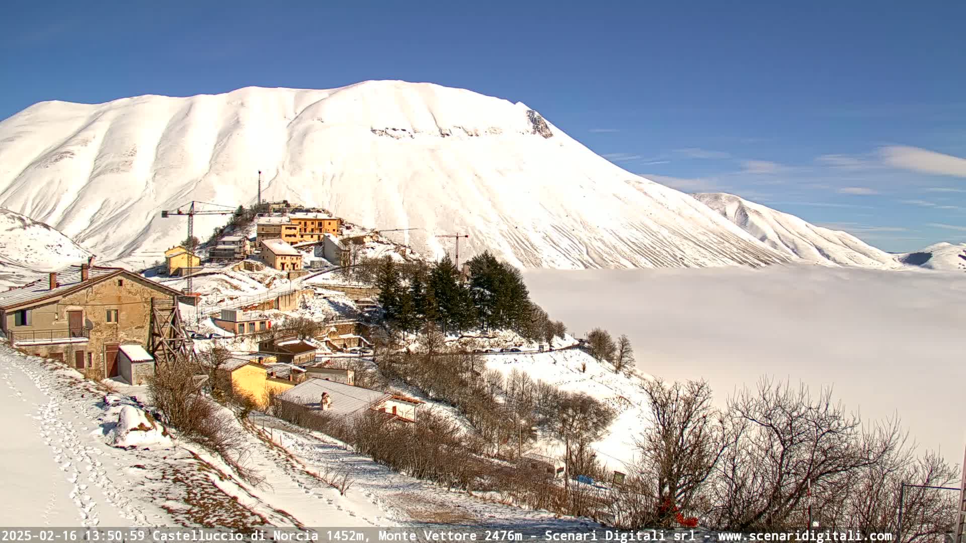 A snow-covered village nestled in a valley below a large, snow-capped mountain, with a layer of fog or clouds in the lower valley under a clear blue sky.