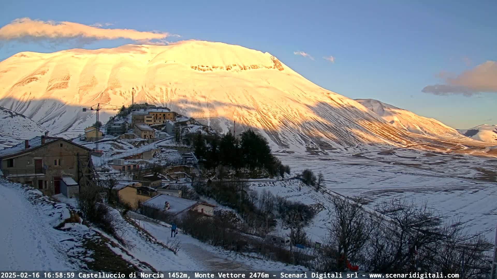 A snow-covered village nestled in a valley at the foot of a large, sunlit, snow-capped mountain under a clear, twilight sky.