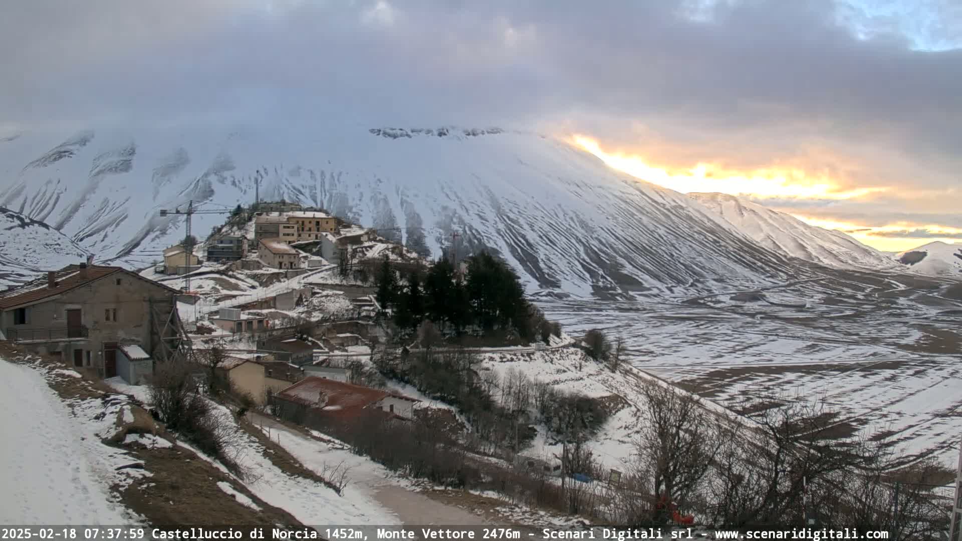 A snow-covered village nestled in a valley, with a large, snow-capped mountain in the background, under a partly cloudy sky with a sunrise or sunset.