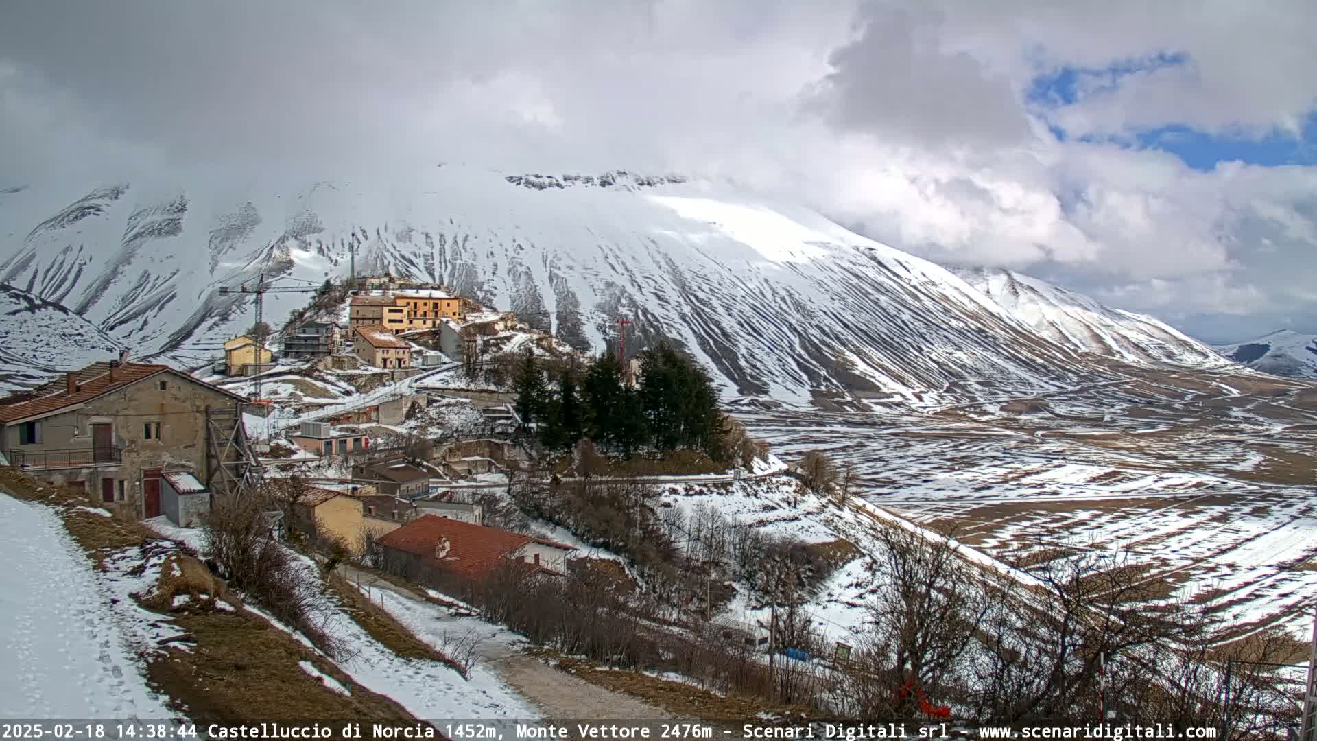 A snow-covered mountainside overlooks a small village nestled in a valley under a cloudy sky.