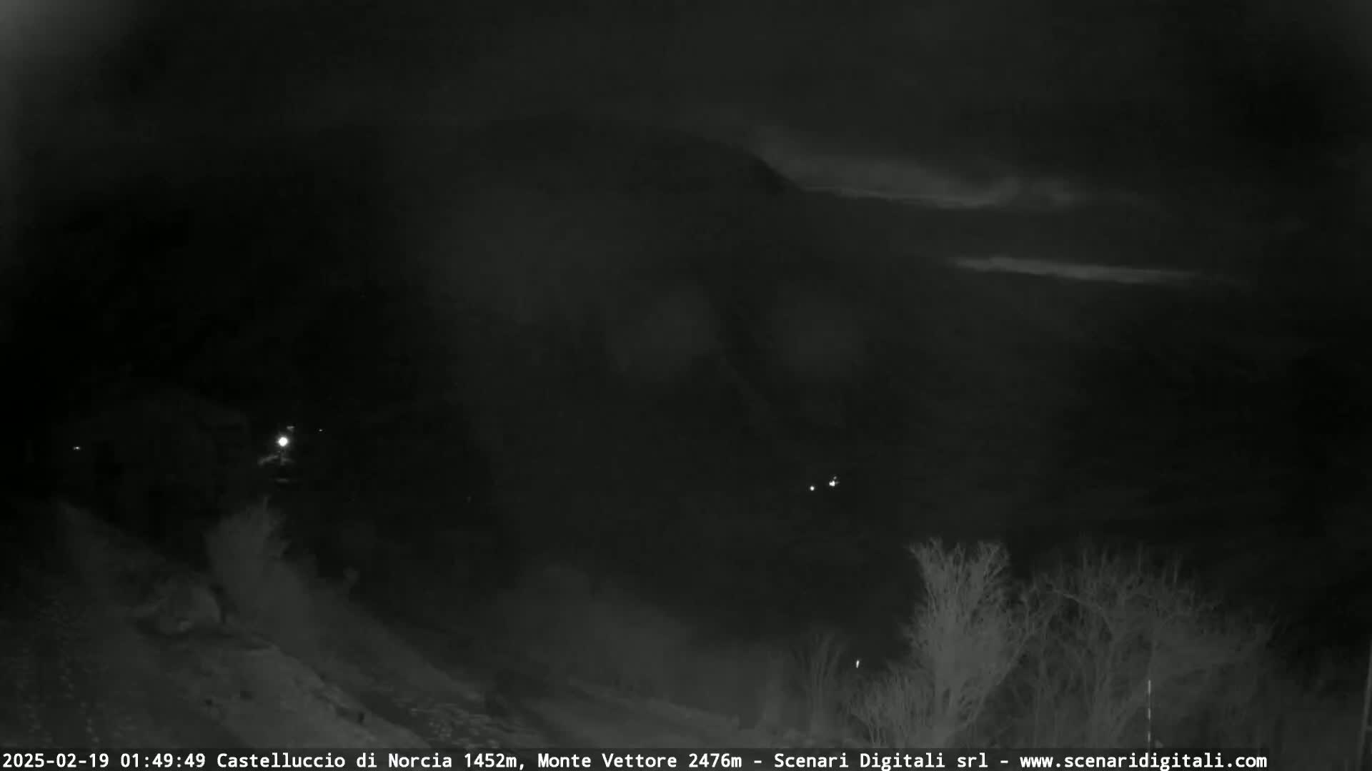 A nighttime, low-light view of a dark, mountainous landscape with a few distant lights and some bare trees in the foreground under a mostly cloudy sky.