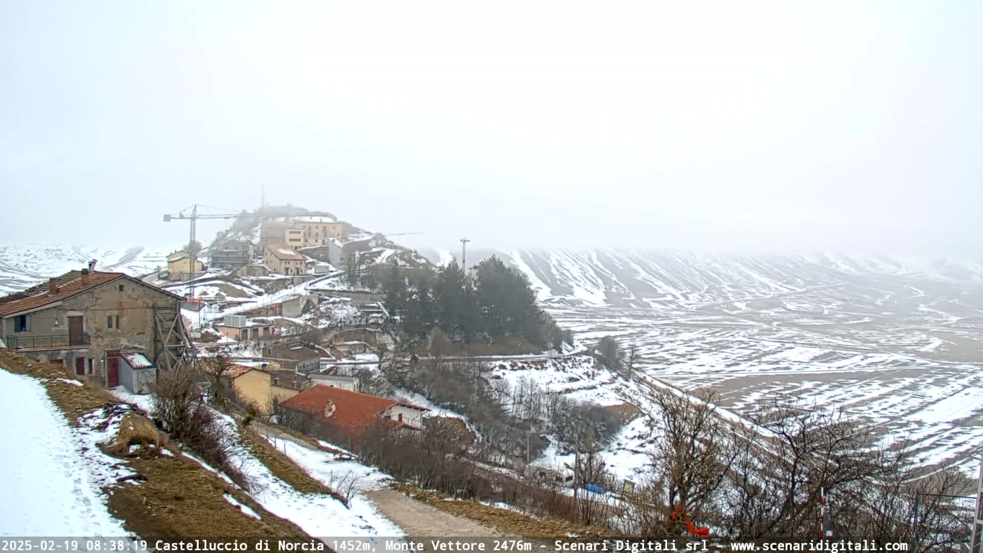 A snow-covered village nestled on a hillside, with a construction crane visible, overlooks a wide, snow-dusted valley under a hazy sky.