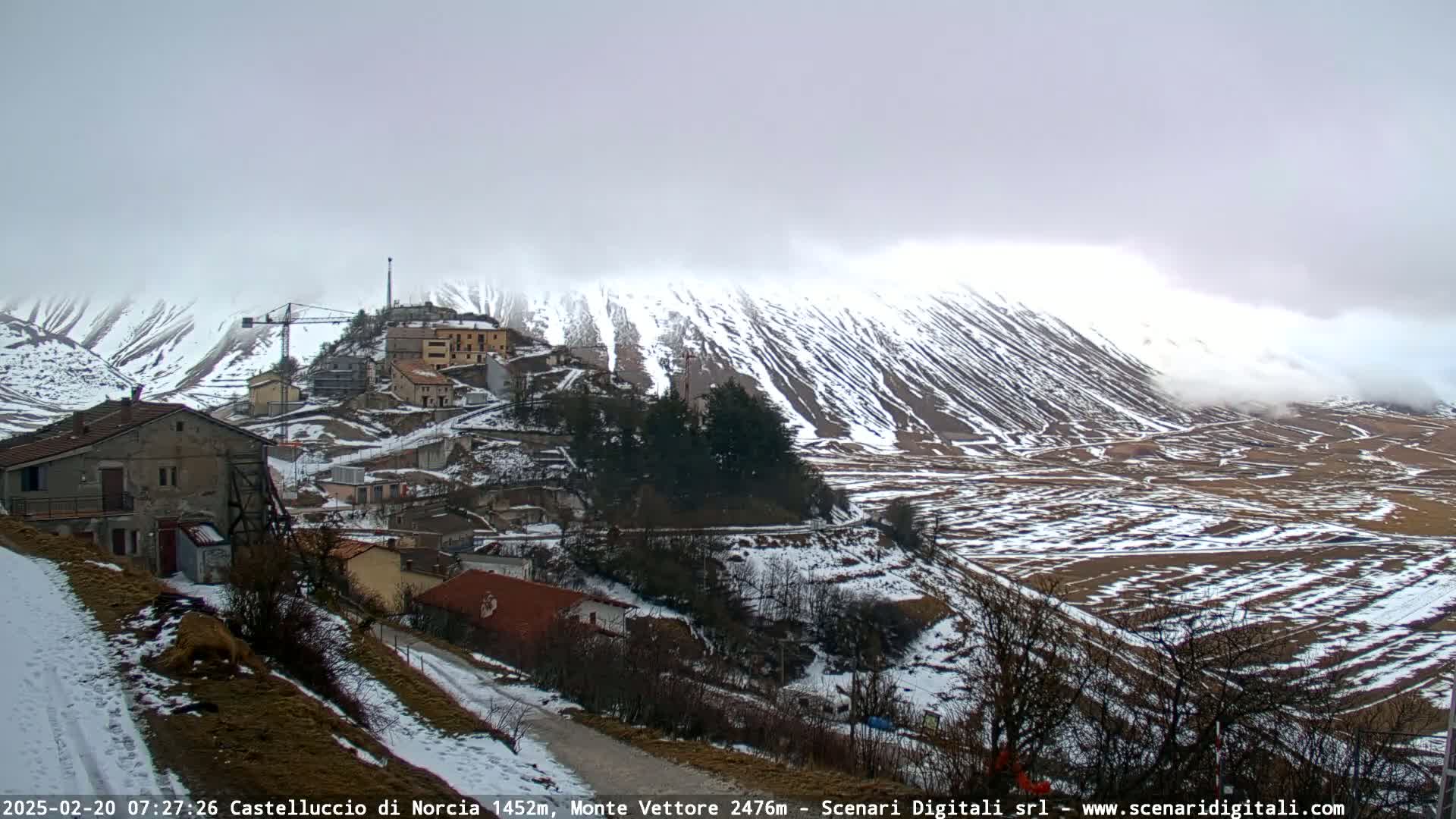A small village sits on a snow-covered hillside beneath a cloudy sky, with a larger snow-covered mountain in the background.