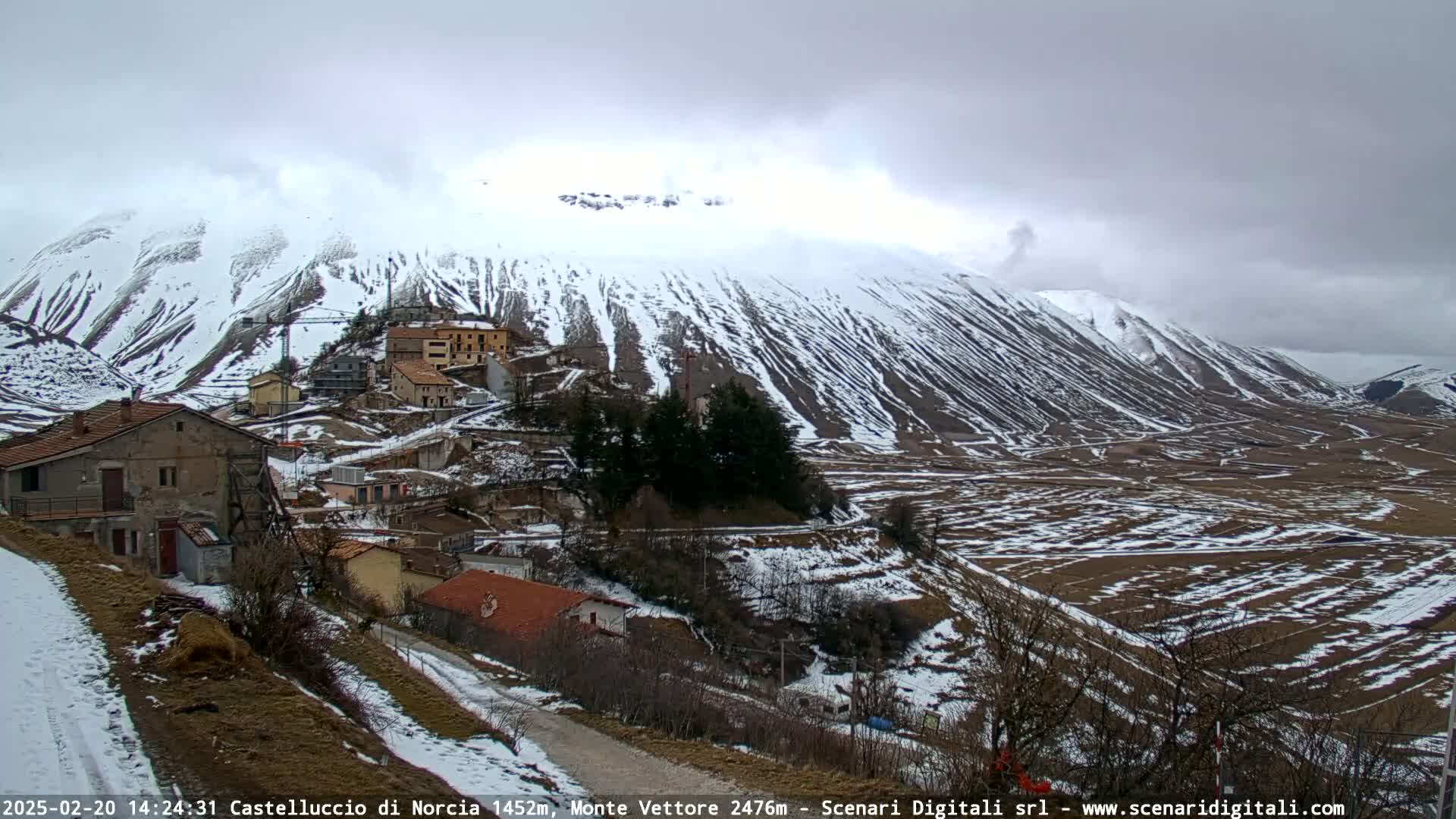 A snow-covered mountain overlooks a small village nestled in a valley under a cloudy sky.