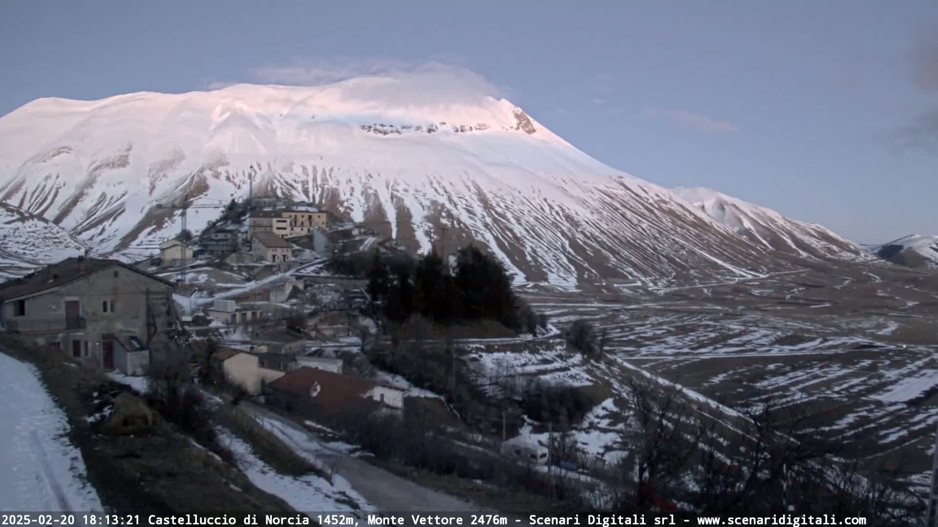 A snow-covered mountain looms over a small village nestled in a valley, under a clear twilight sky.