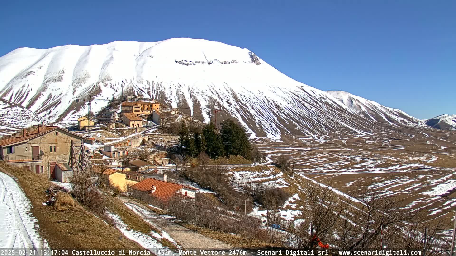 A snow-capped mountain overlooks a small village nestled in a valley on a sunny day.