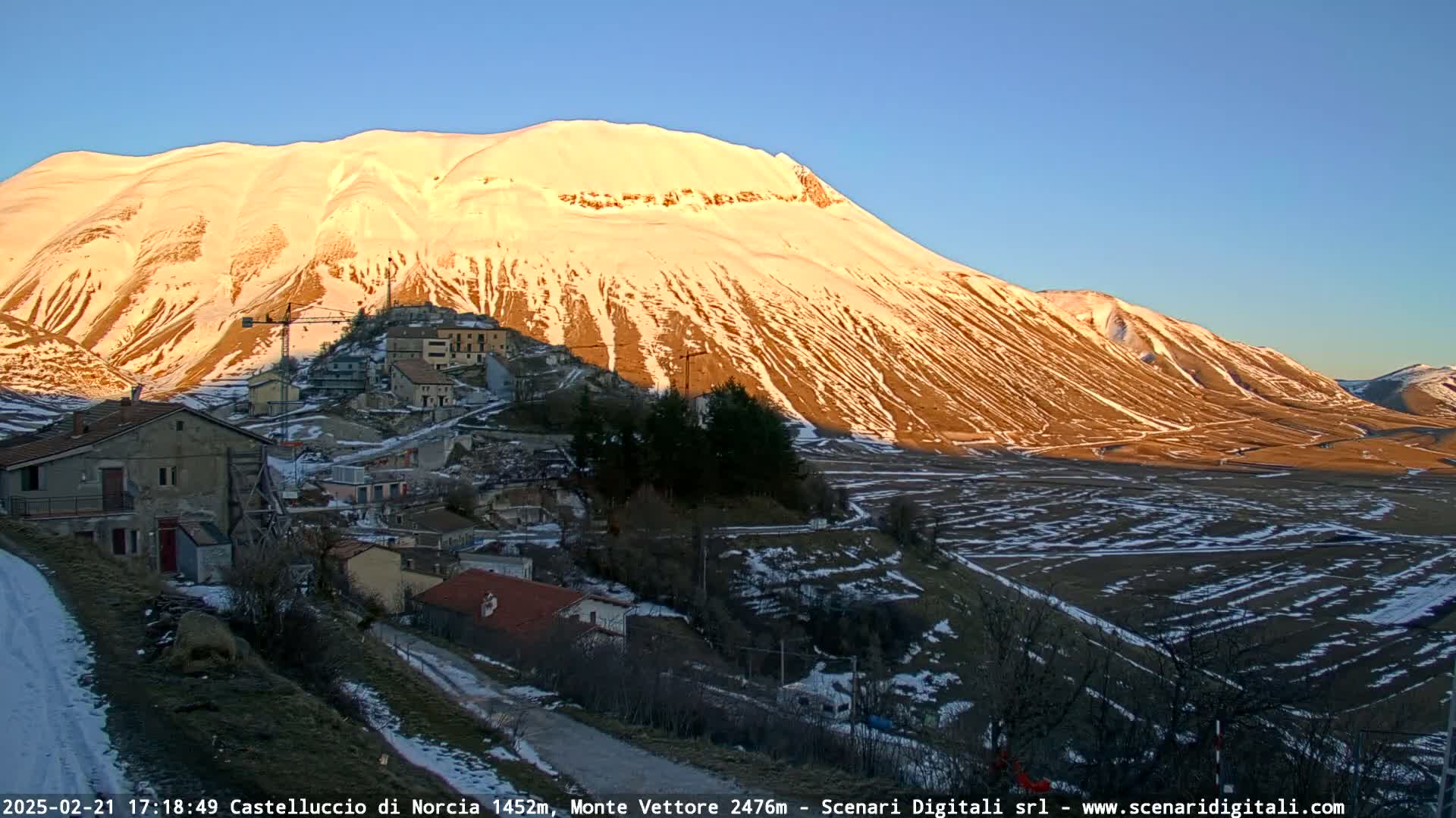 A snow-dusted mountainside is illuminated by the setting sun, overlooking a small village nestled in a valley with patches of snow on the ground.