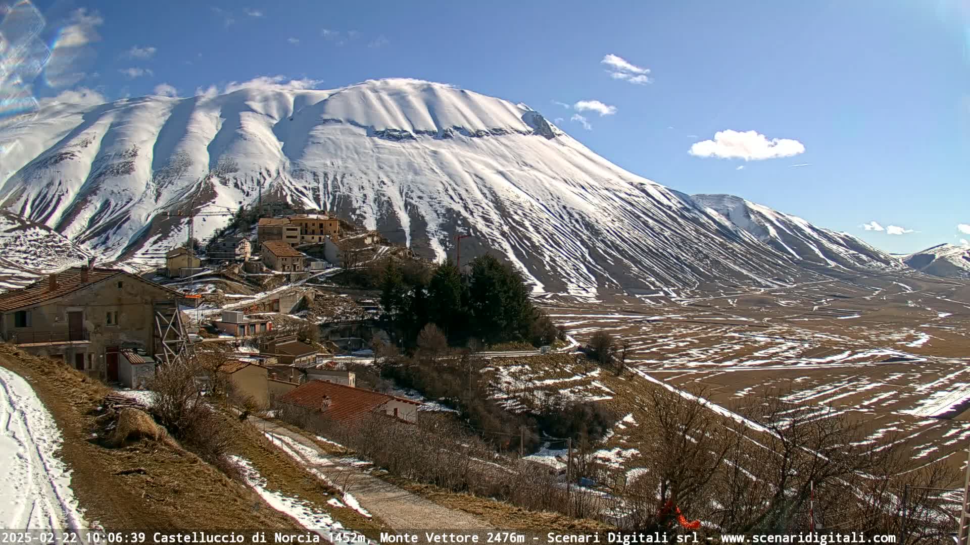 A snow-covered mountain looms over a small village nestled in a valley on a sunny day.