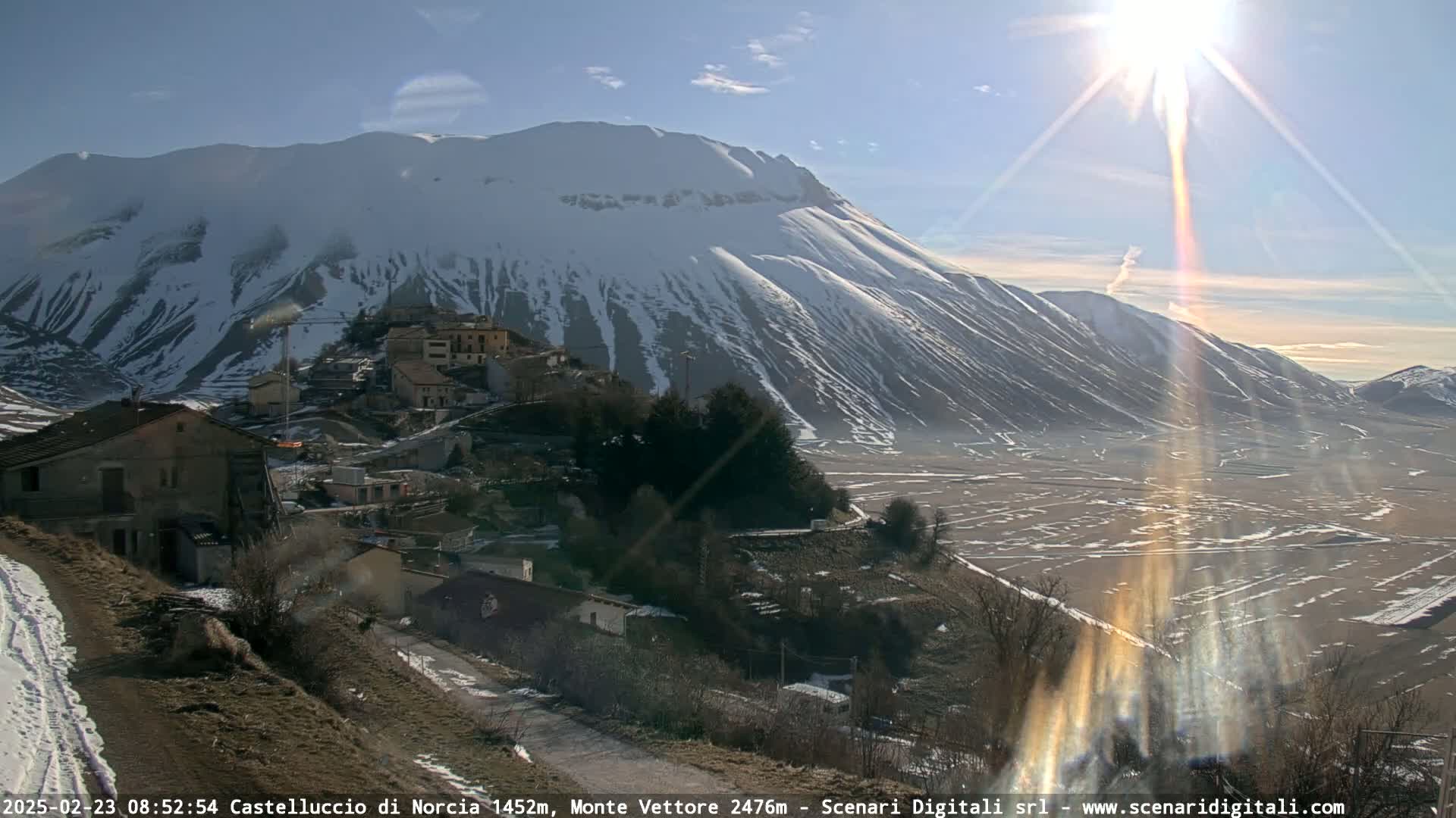 A snow-capped mountain looms over a small village nestled in a valley on a sunny day.