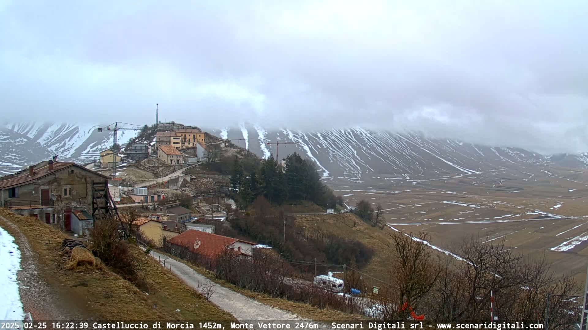 A small village nestled in a valley, partially snow-covered, with construction cranes visible and a mountain range in the background under a cloudy, overcast sky.