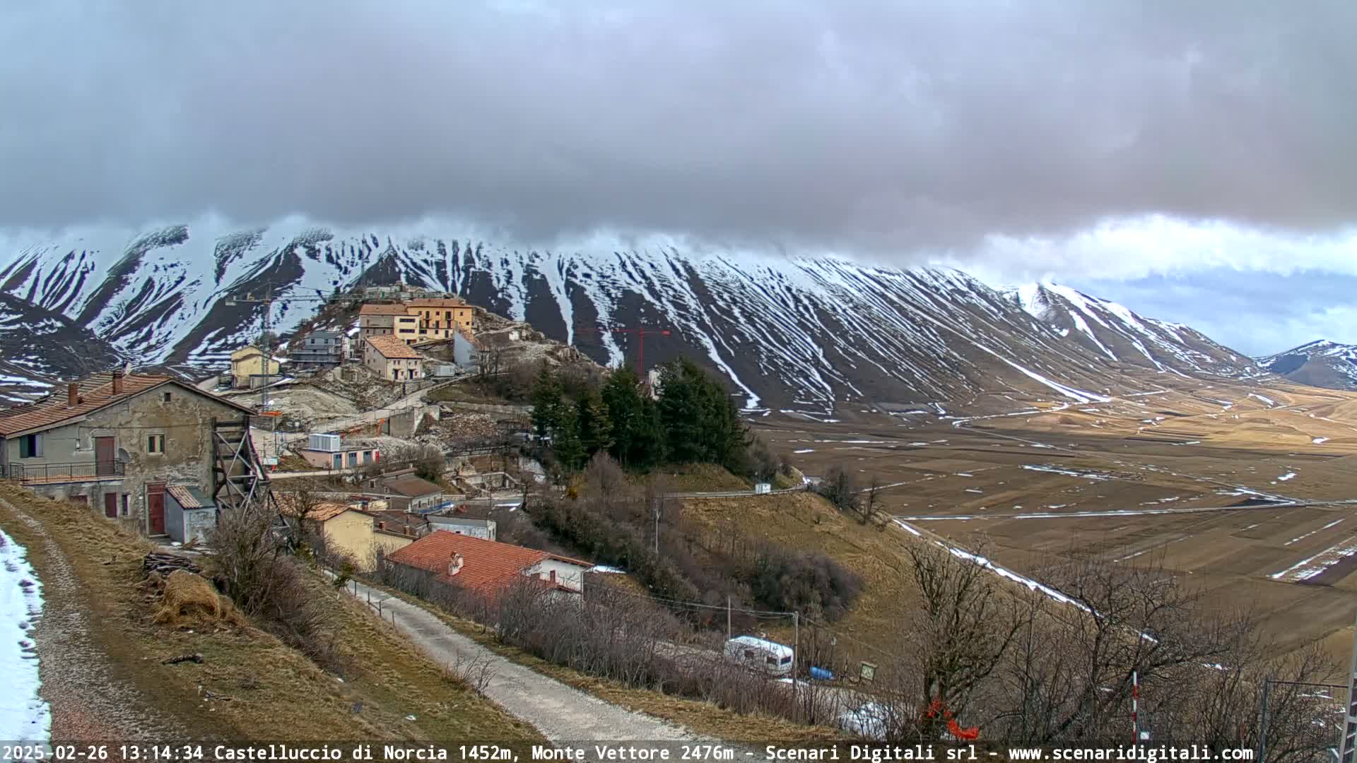 A small village nestled in a valley, with snow-dusted mountains under a cloudy sky.