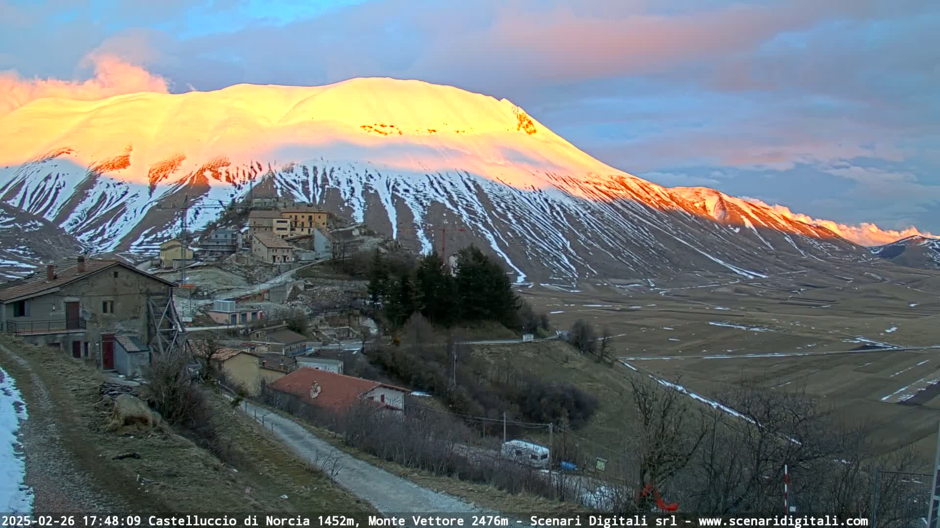 A snow-capped mountain glows orange in the sunset light, overlooking a small village nestled in a valley.