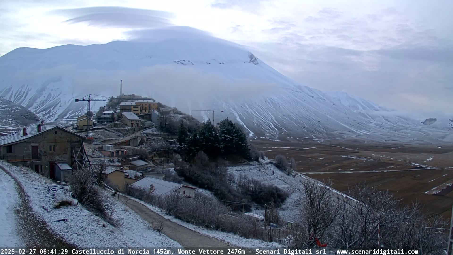 A snow-covered village nestled in a valley, with a large, snow-capped mountain in the background under an overcast sky.