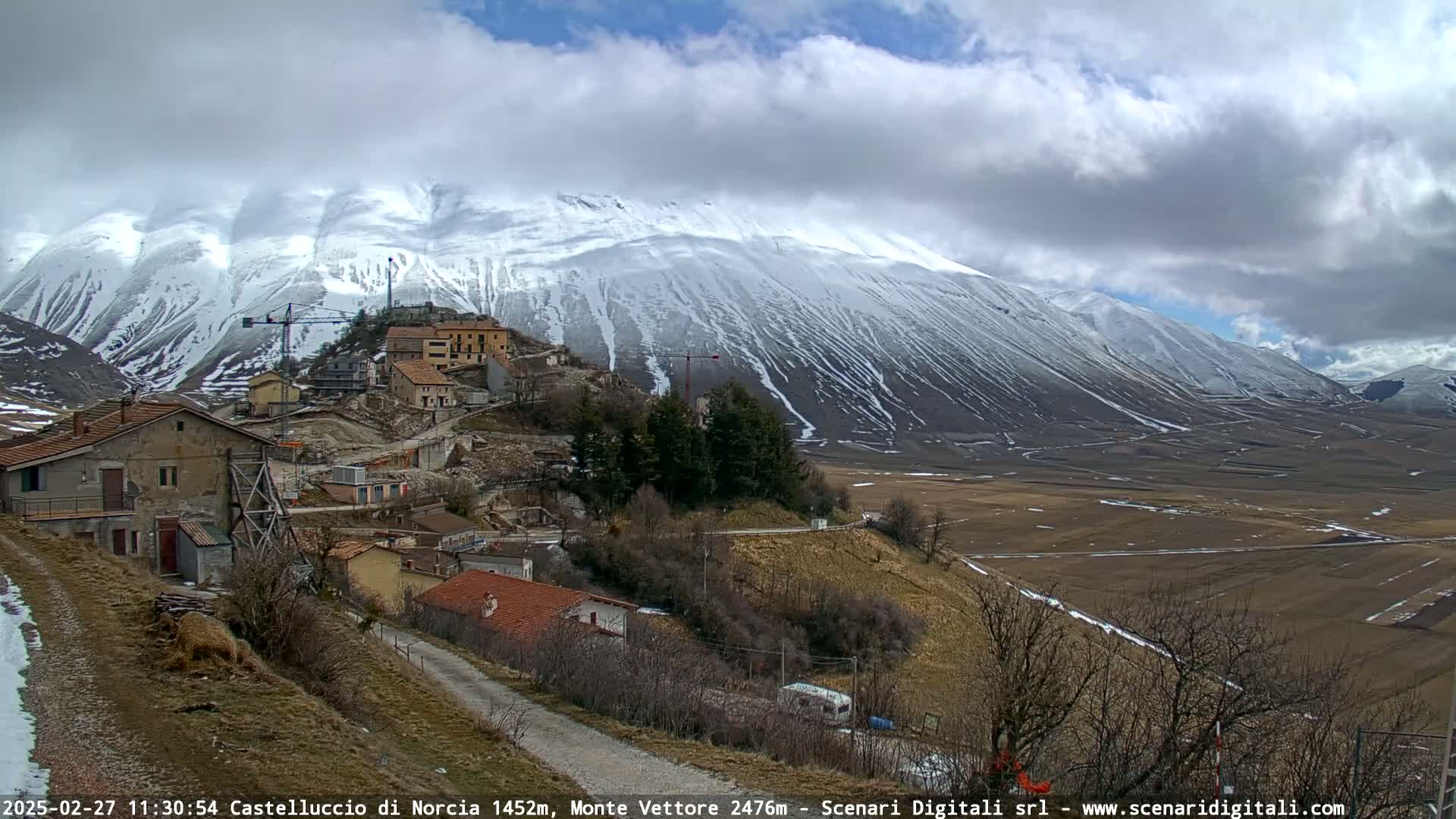 A small village nestled on a hillside below a large, snow-capped mountain range under a partly cloudy sky.