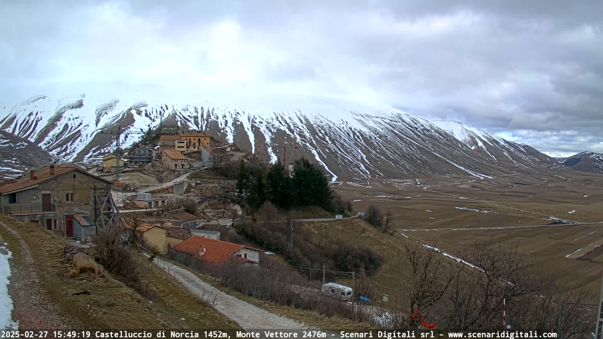 A snow-dusted mountain overlooks a small village nestled in a valley under a cloudy sky.