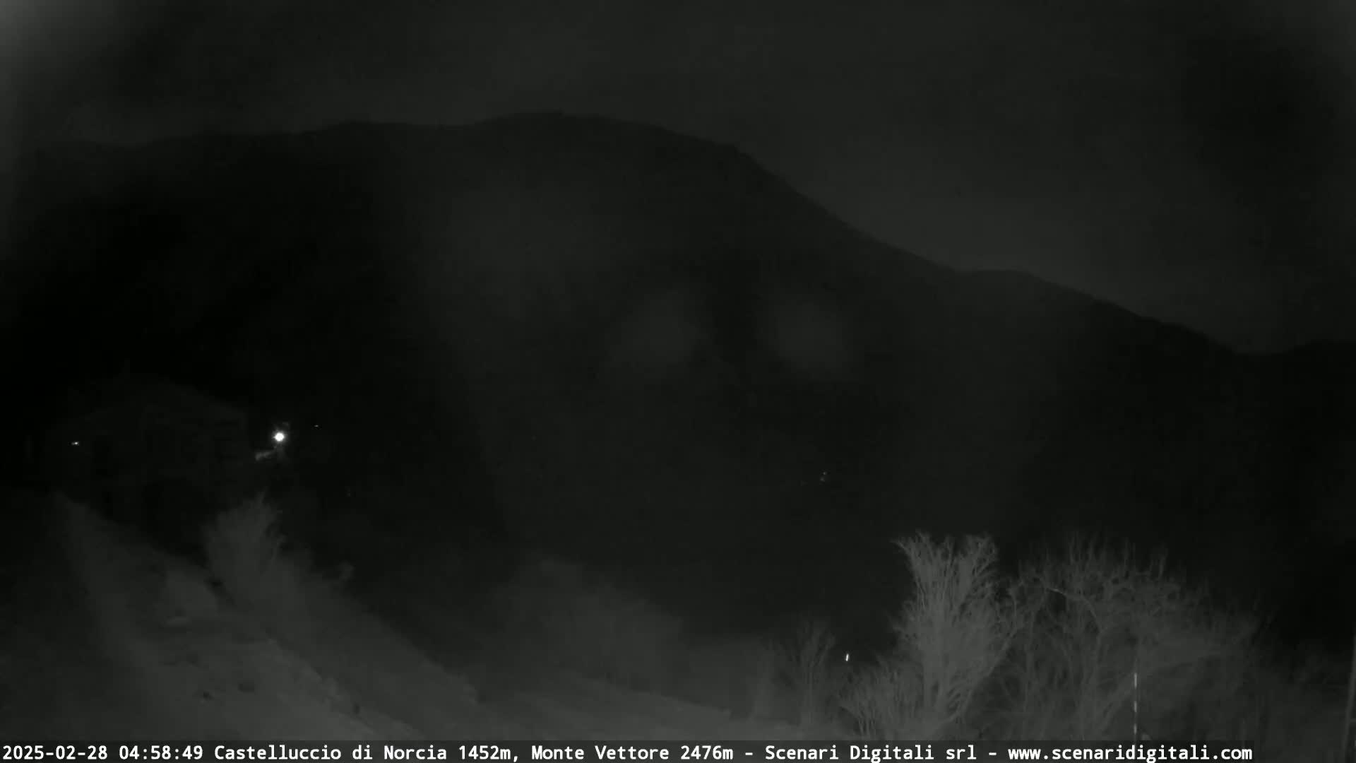 A dark, nighttime view of a mountain silhouetted against a mostly clear sky, with some sparse, leafless trees in the foreground.