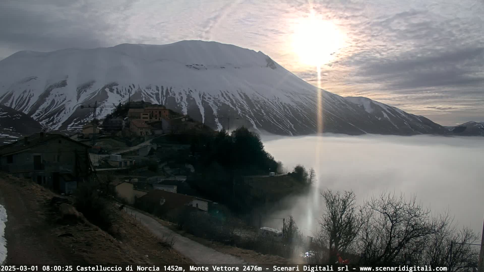 A snow-capped mountain at sunrise casts a sunbeam over a fog-covered valley containing a small village.