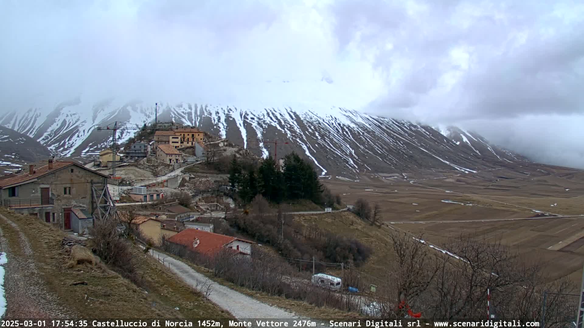 A small village nestled in a valley, with a snow-dusted mountain in the background under a cloudy sky.