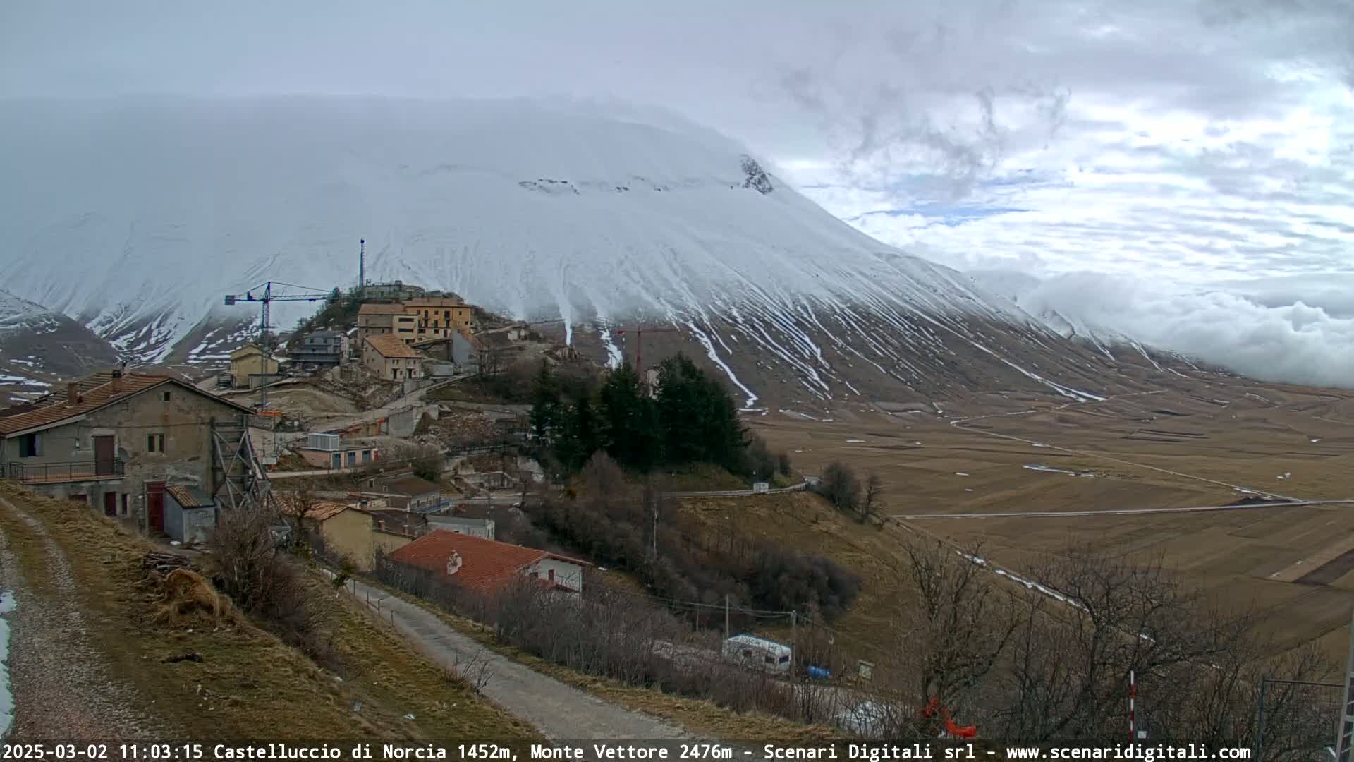 A snow-covered mountain looms over a small village nestled in a valley under a cloudy sky.