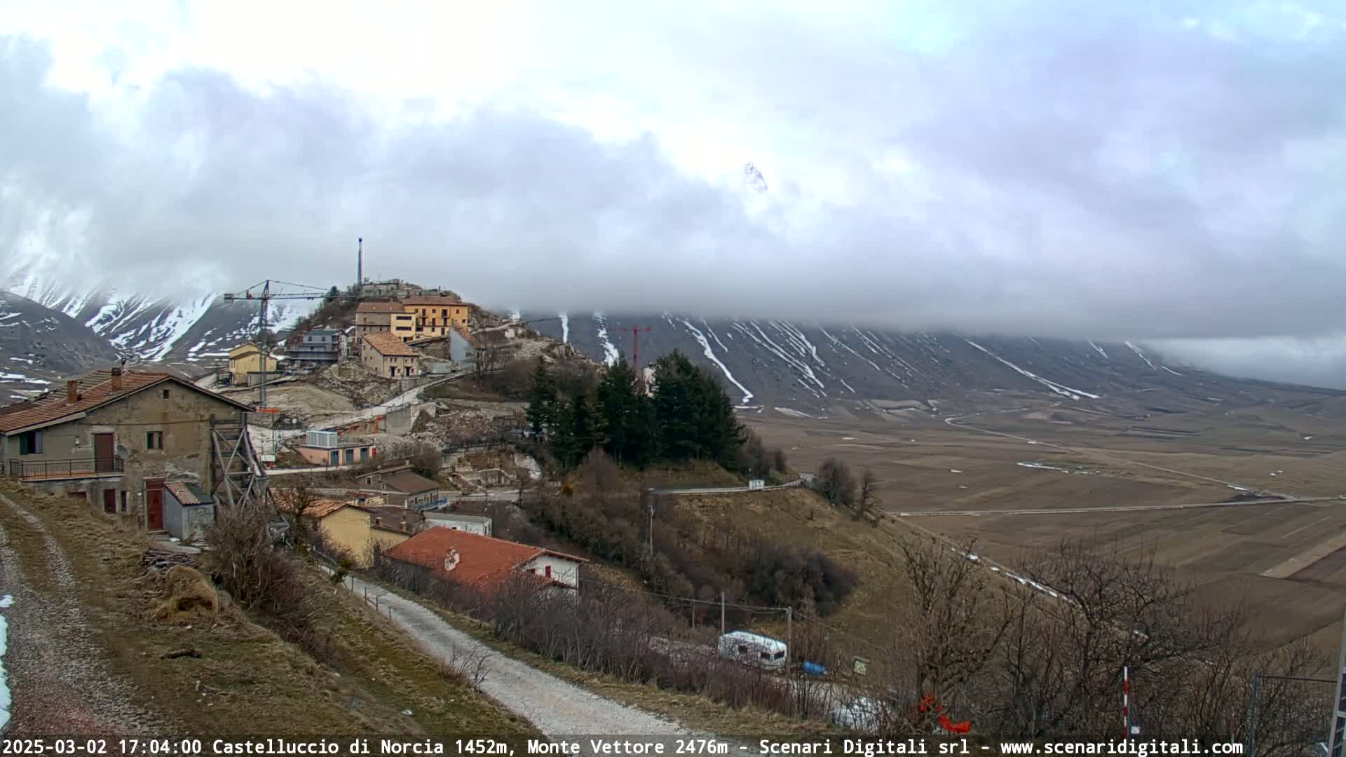 A small village nestled on a hillside, with a partially cloudy sky and snow-dusted mountains in the background.