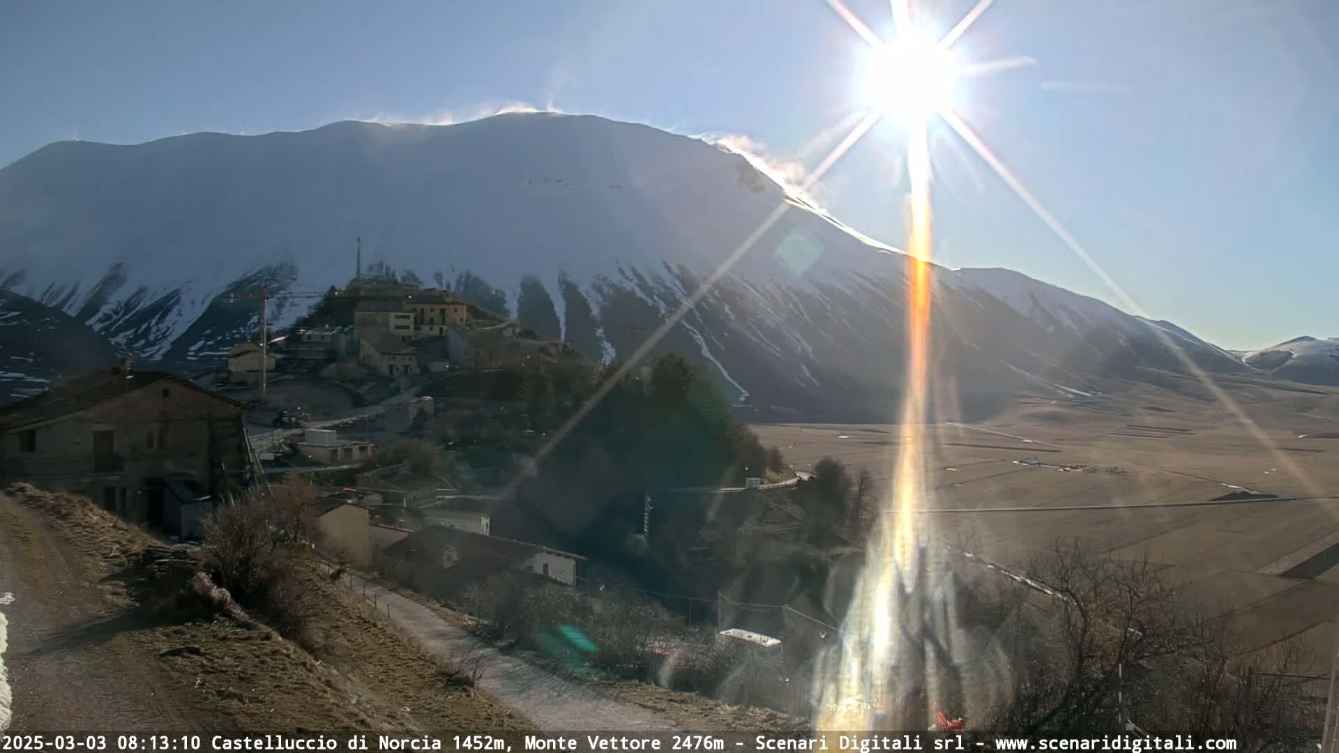 A sunlit village nestled below a snow-capped mountain on a clear, bright day.