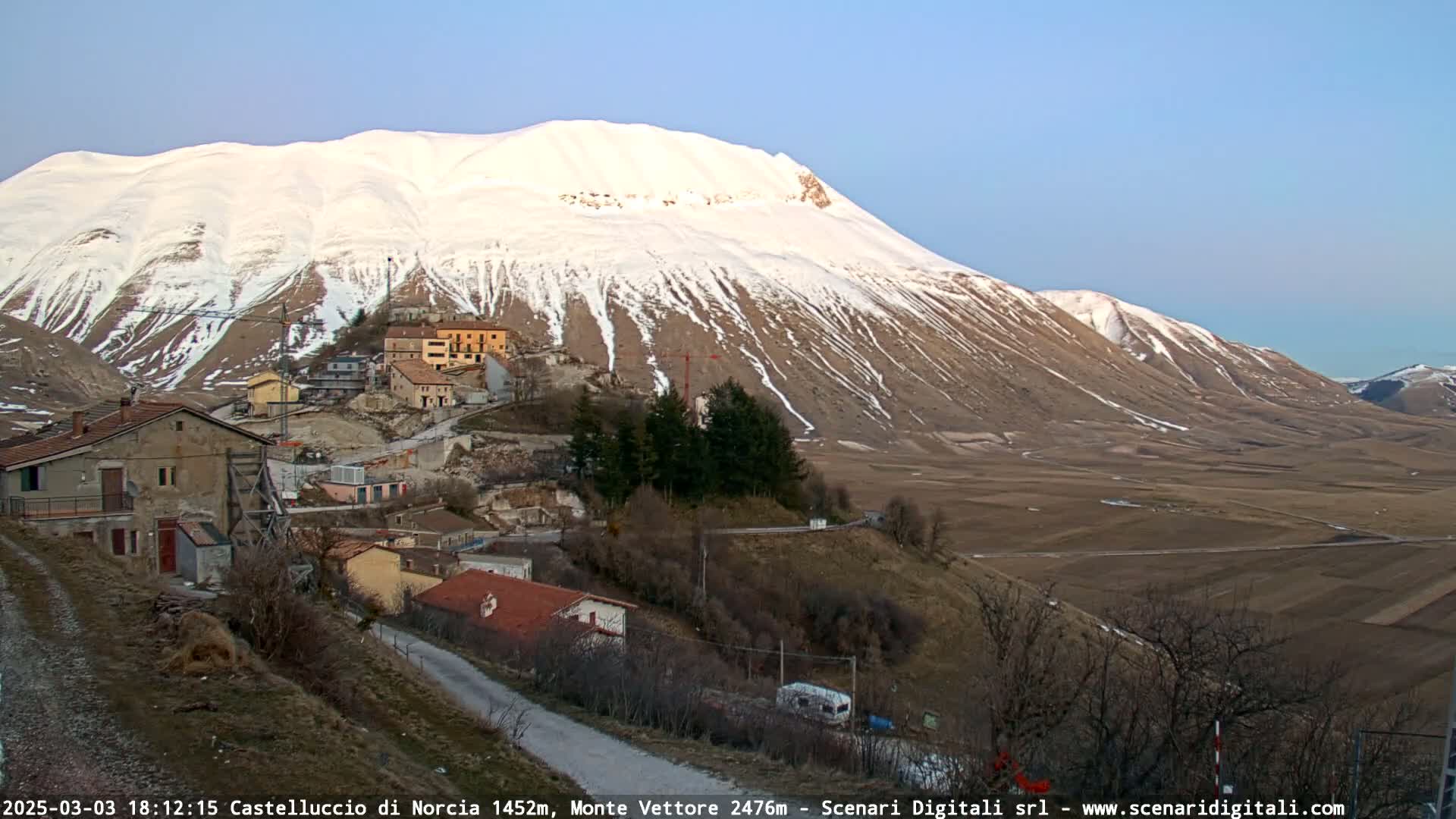 A snow-capped mountain overlooks a small village nestled in a valley, under a clear, pale blue sky.