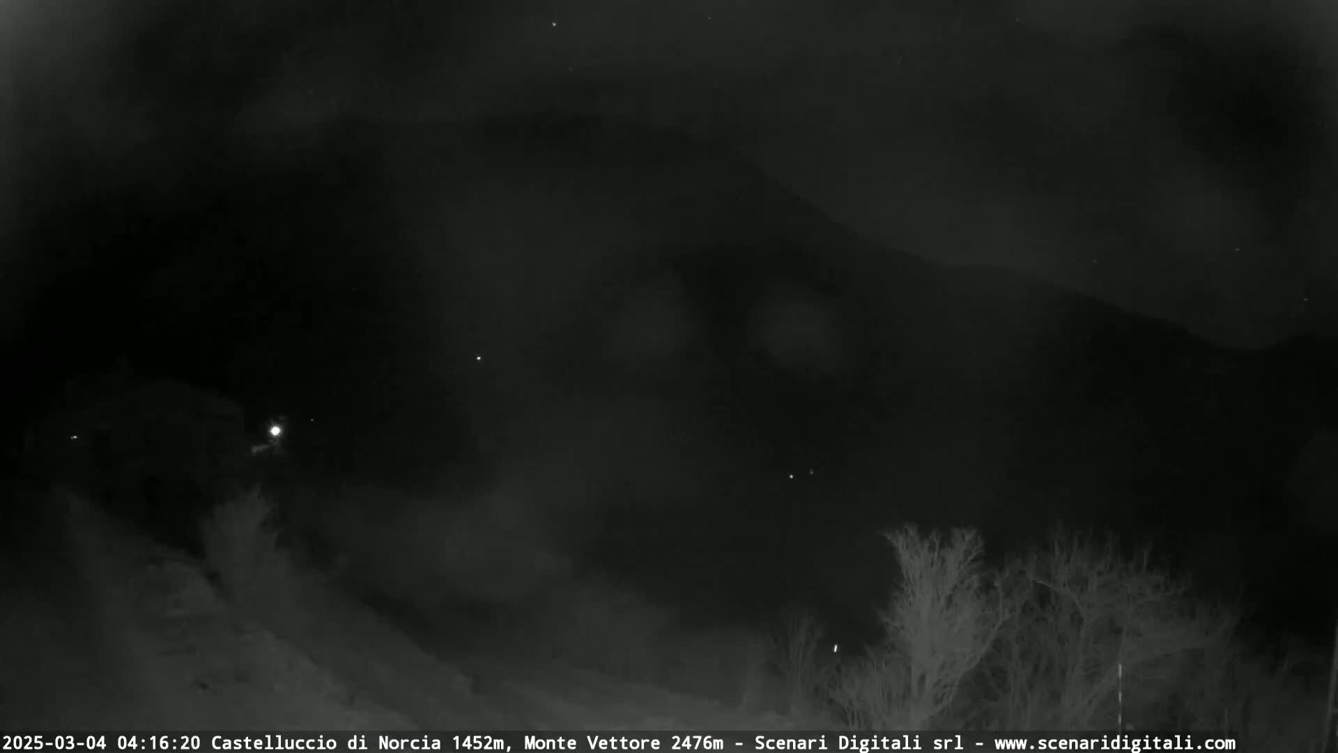 A nighttime, low-light view shows a dark mountain against a mostly clear sky with a few faint lights visible in the distance and some bare trees in the foreground.