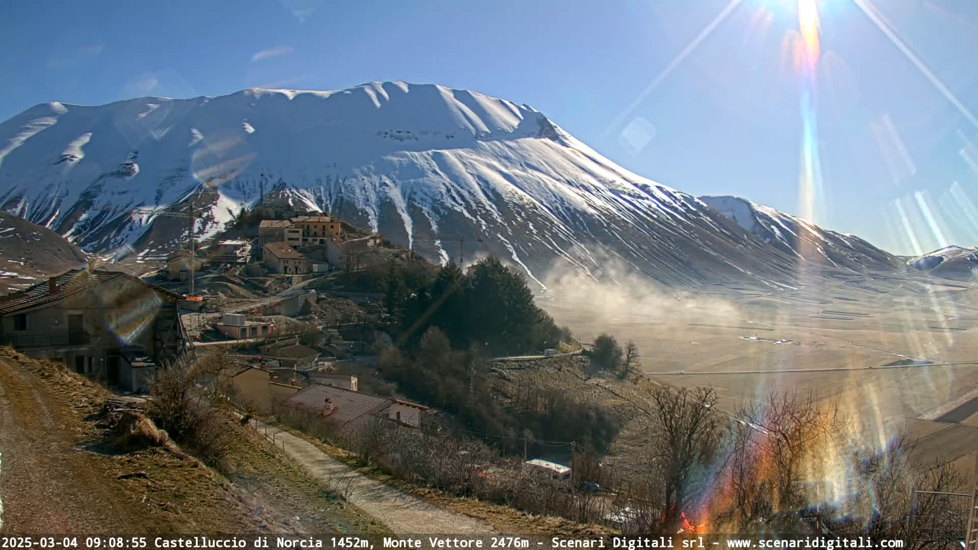 A snow-capped mountain overlooks a small village nestled in a valley on a sunny day with a light haze in the distance.