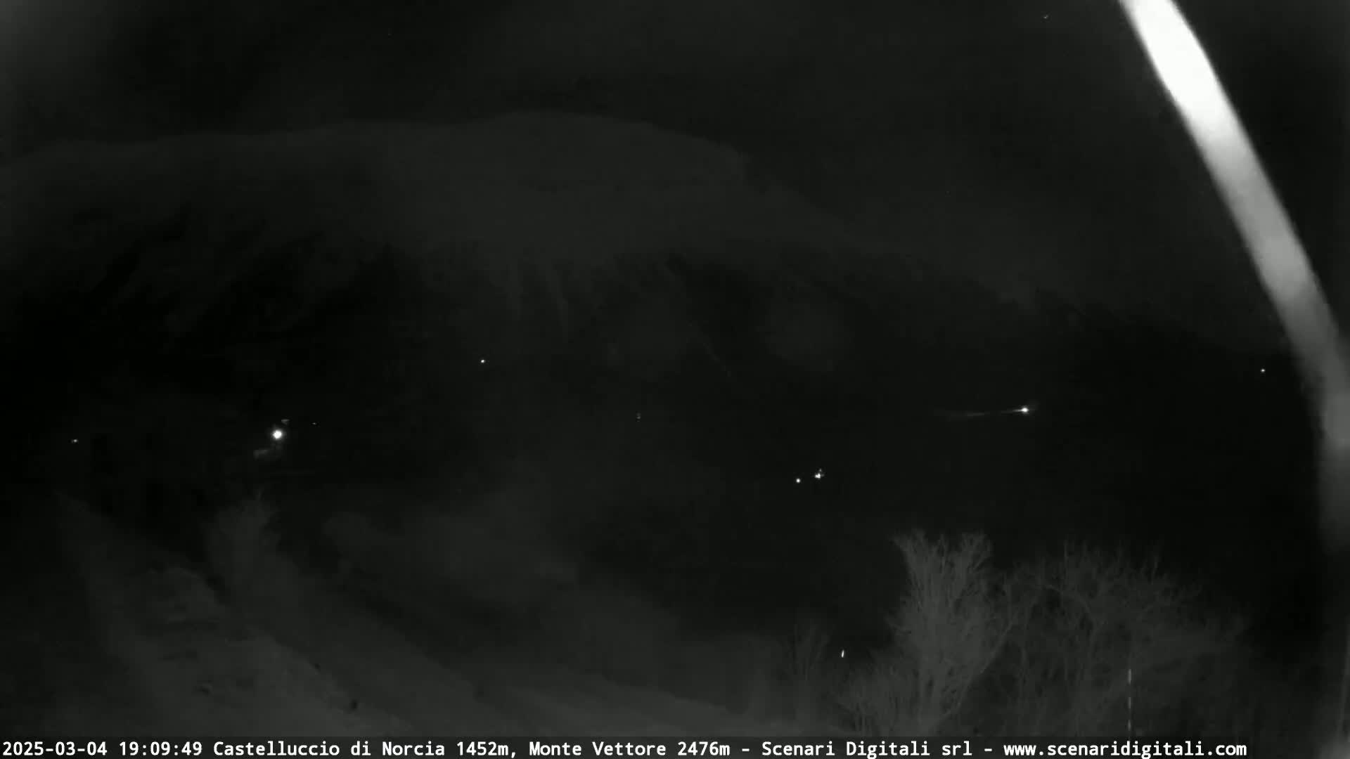 A nighttime, low-light view of a snow-covered landscape with a few distant lights visible under a dark, cloudy sky.