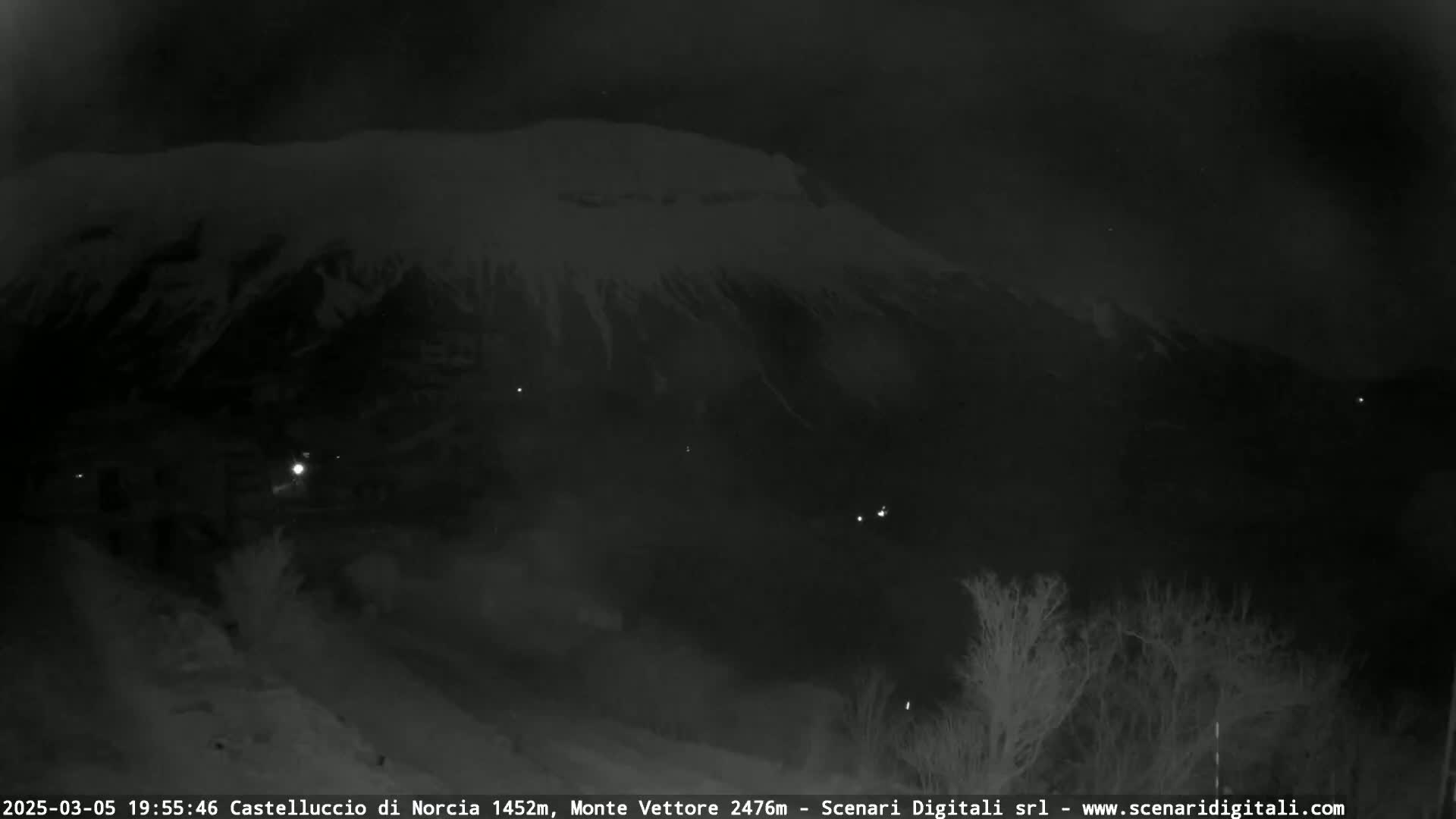 A snow-covered mountain is seen at night, with a few faint lights visible in the foreground and the middle ground.