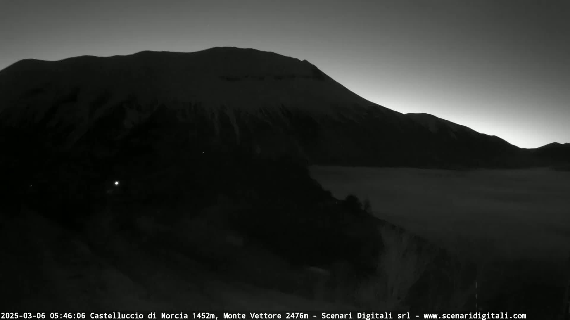 A dark, snow-covered mountain range is silhouetted against a pre-dawn sky, with a low-lying layer of fog or clouds in the valley below.