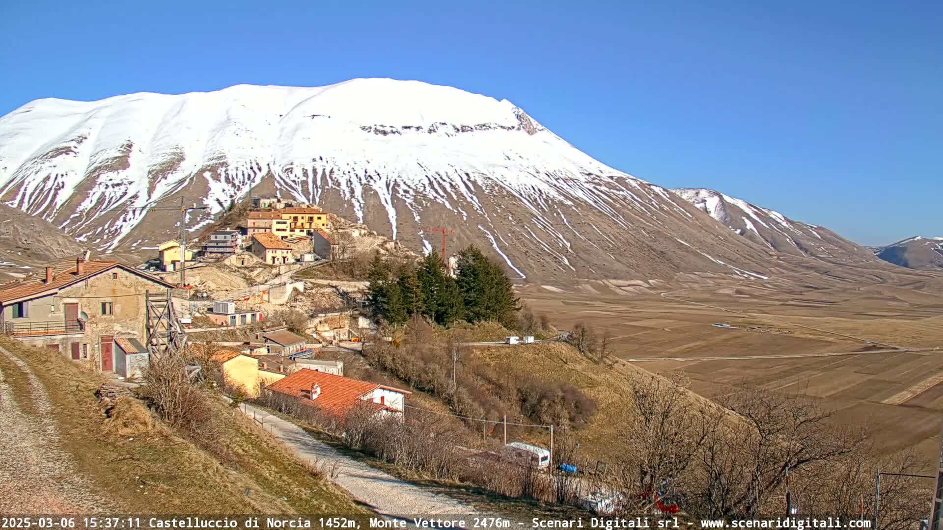 A small village nestled on a hillside below a snow-capped mountain range on a sunny day.