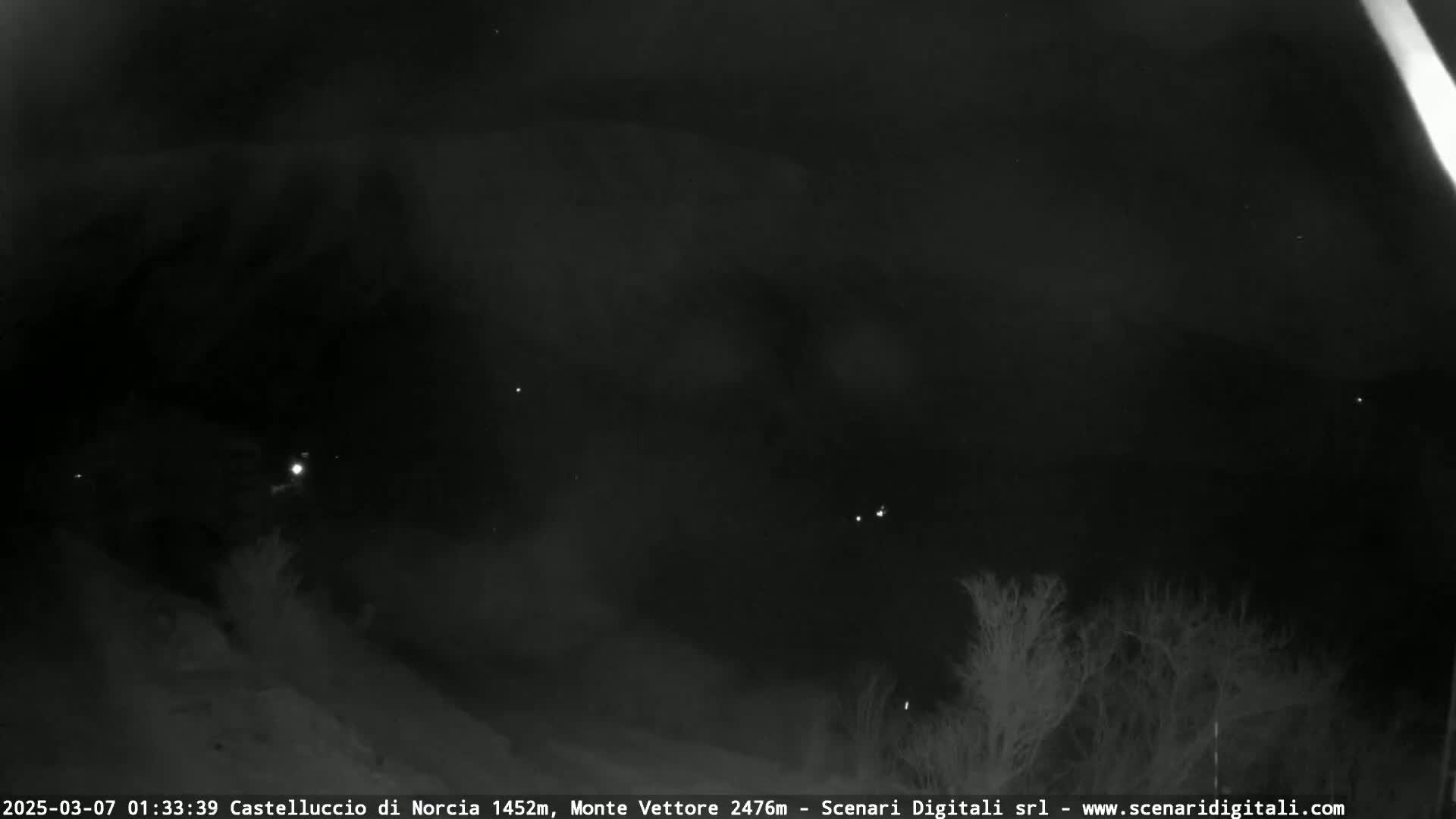 A nighttime outdoor scene shows a dark, cloudy sky with a few faint lights visible in the distance, and some silhouetted trees in the foreground.