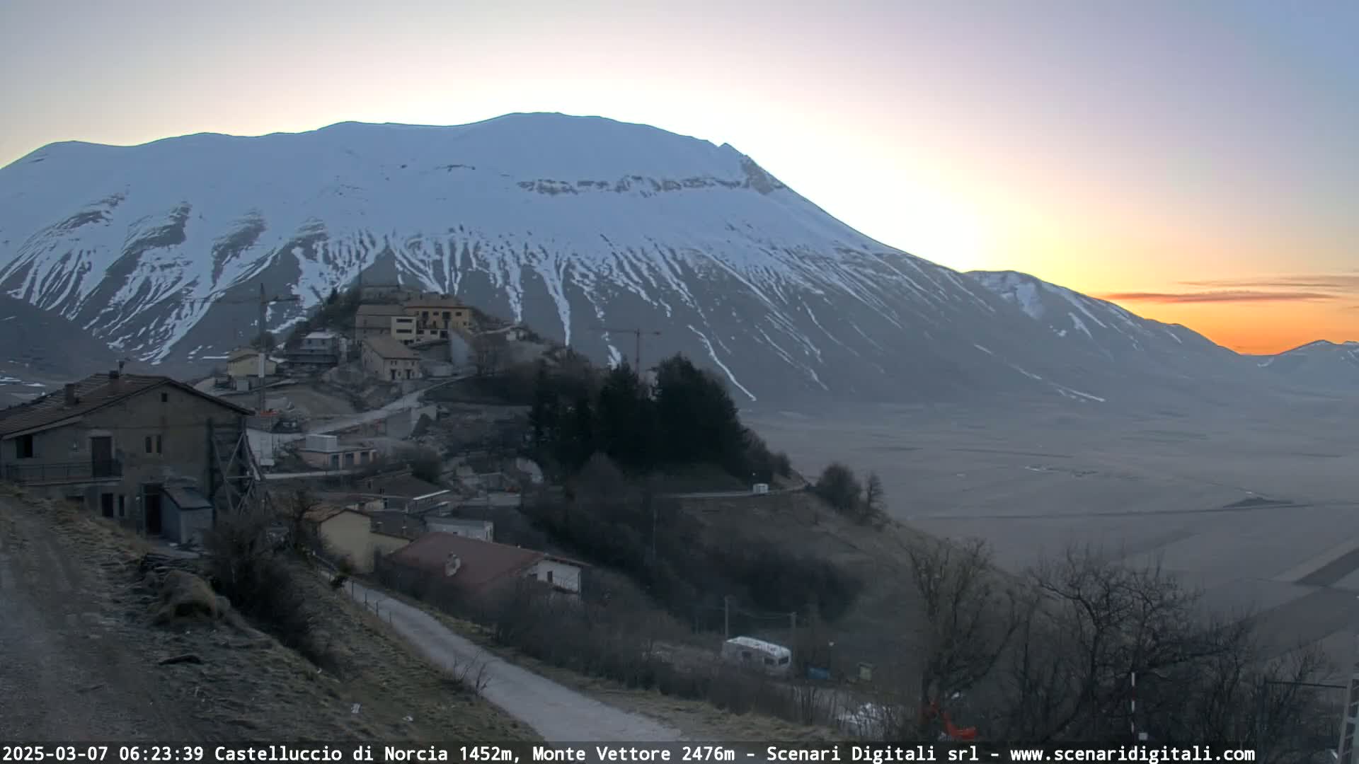 A small village nestled on a hillside at sunrise, with a large snow-capped mountain in the background.