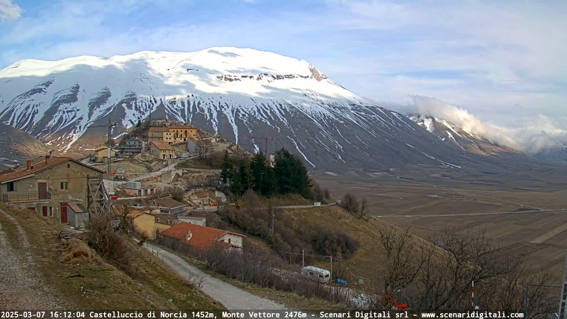 A small village nestled on a hillside overlooks a vast, flat valley and a snow-capped mountain range under a mostly sunny sky with some clouds.