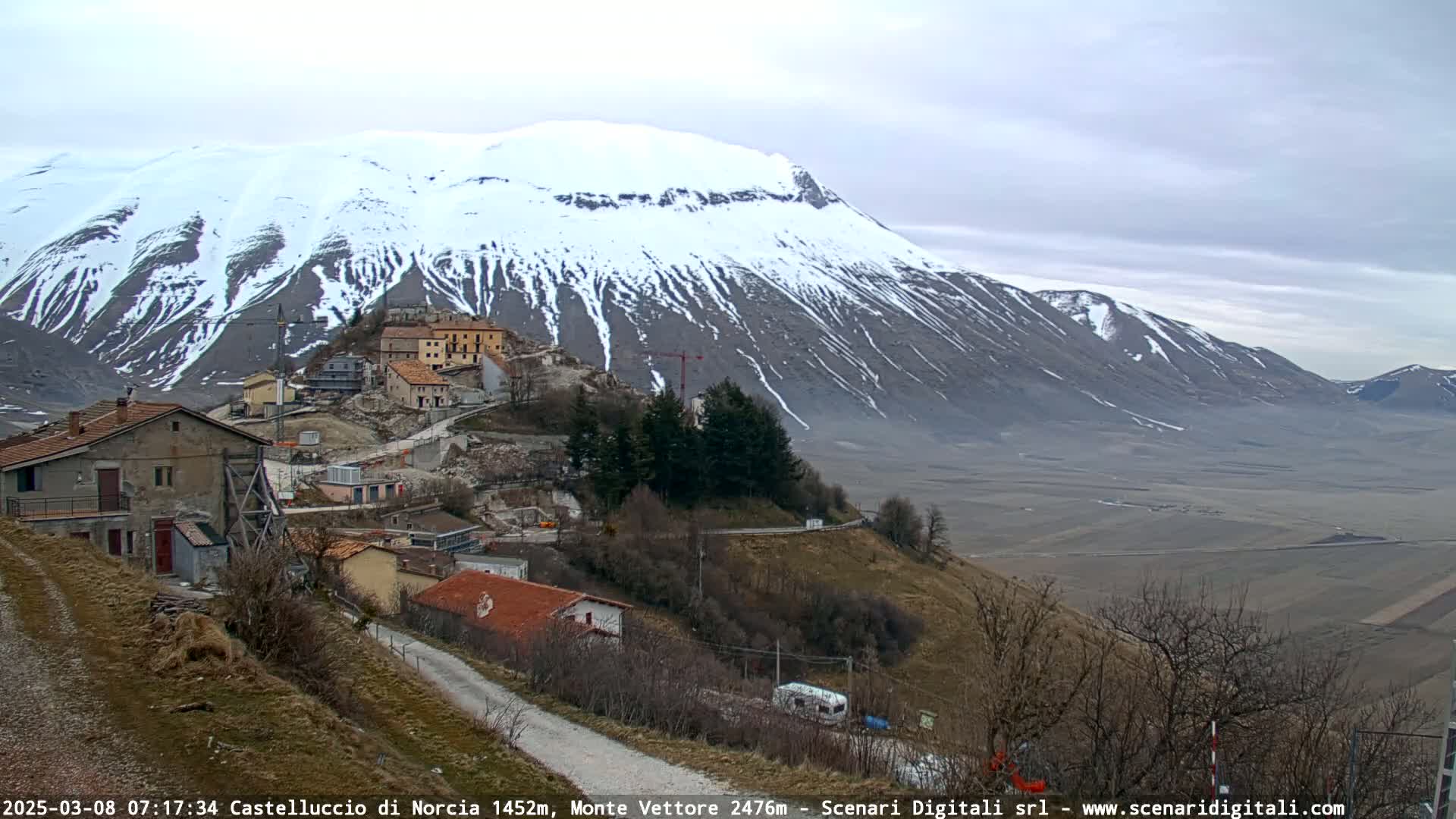A snow-capped mountain overlooks a small village nestled on a hillside, under an overcast sky.