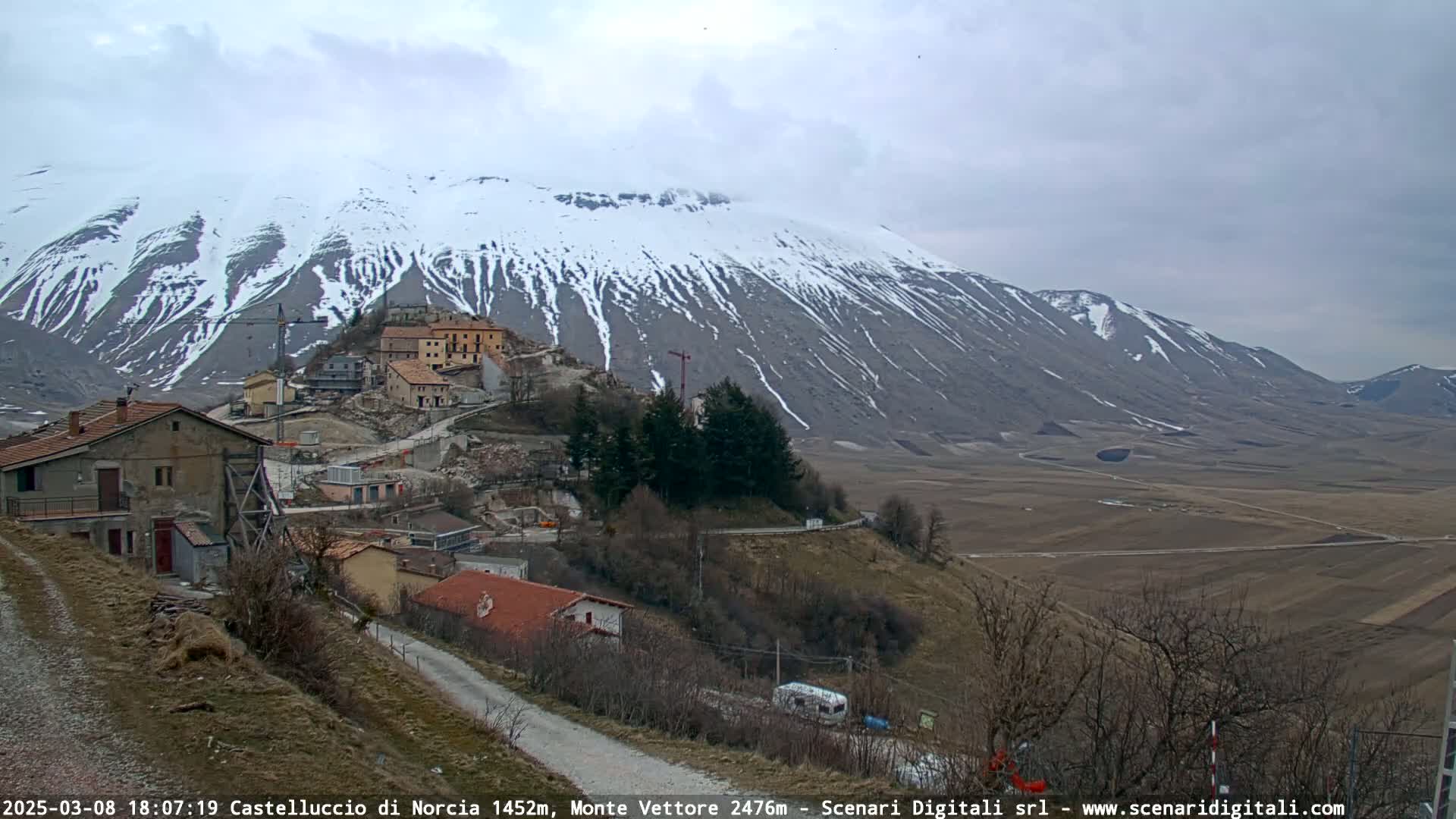 A snow-capped mountain overlooks a small village nestled in a valley under an overcast sky.