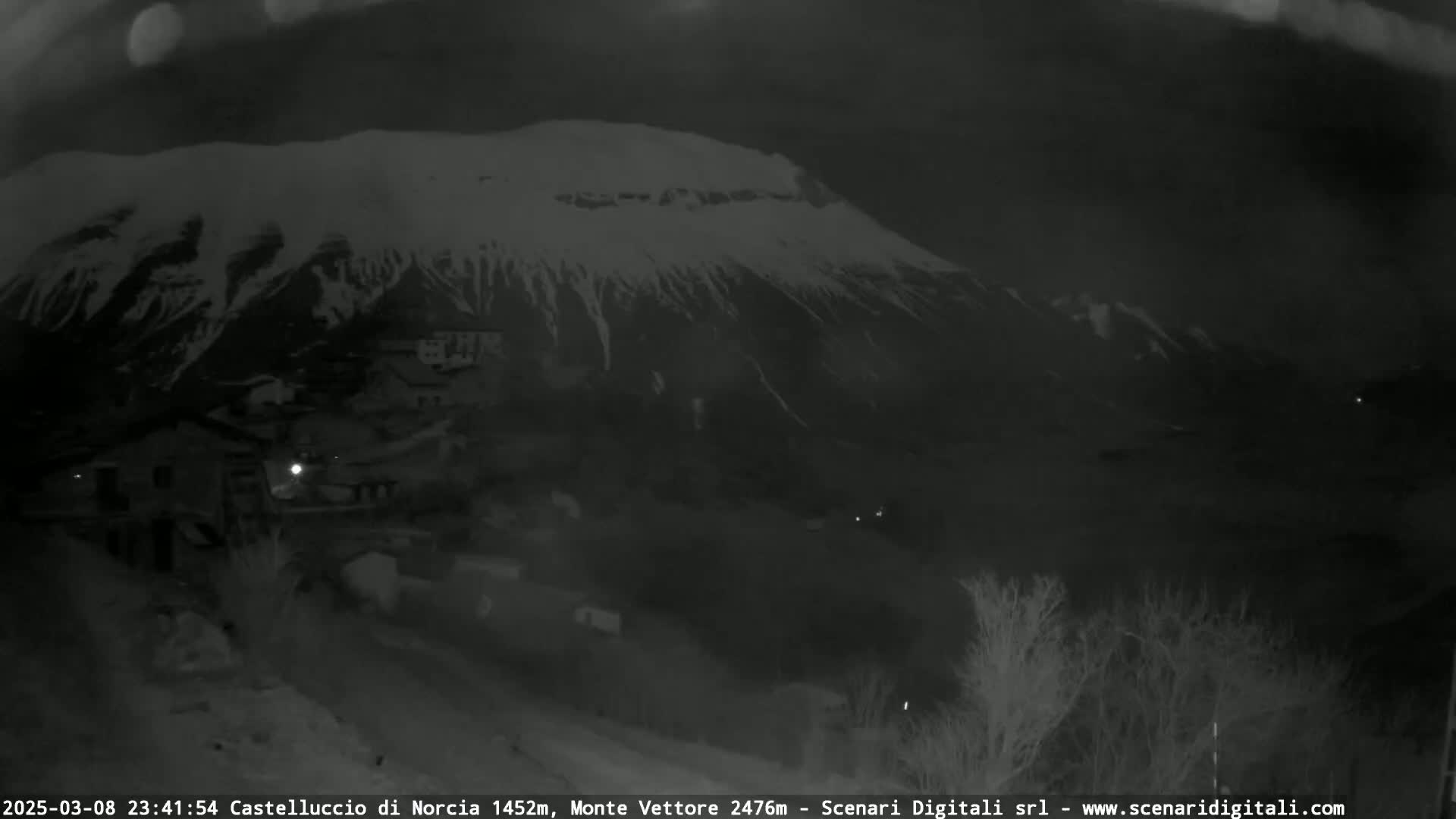 A nighttime, low-light view shows a snow-capped mountain looming over a small village under a dark sky.