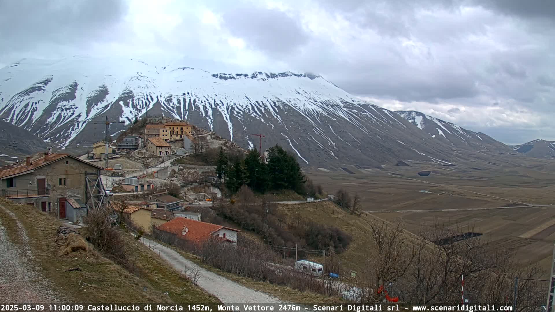 A snow-capped mountain overlooks a small village nestled in a valley under a cloudy sky.