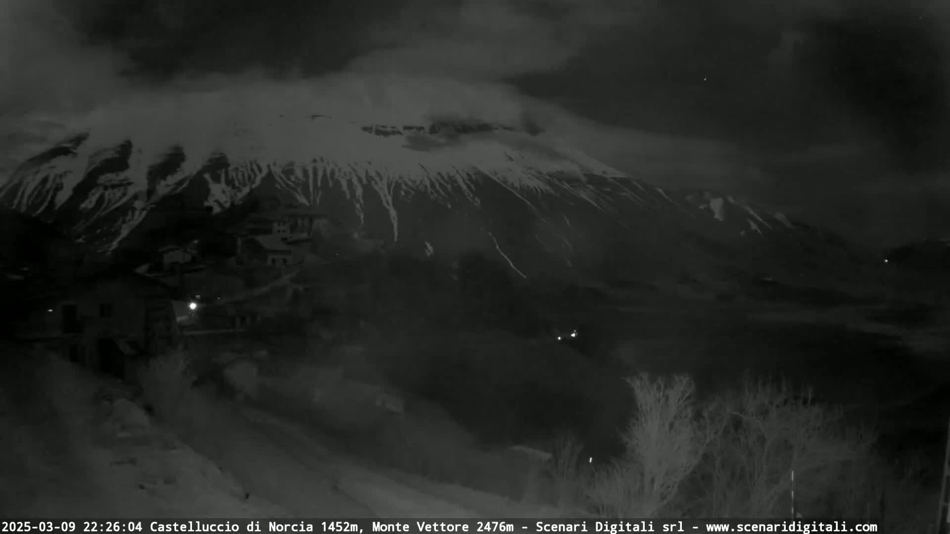A nighttime, overcast view shows a snow-covered mountain range behind a small village.