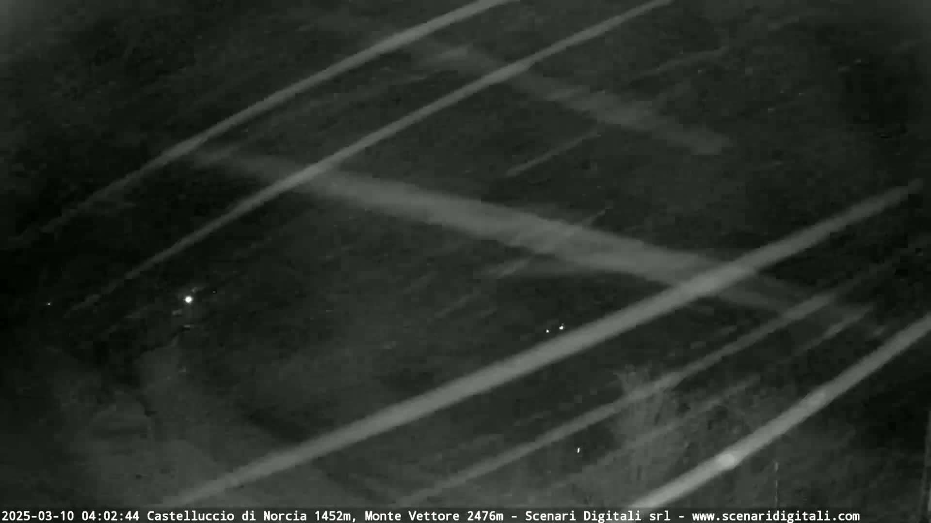 A nighttime, snowy scene is shown with diagonal streaks of light possibly from car headlights cutting through the snow.